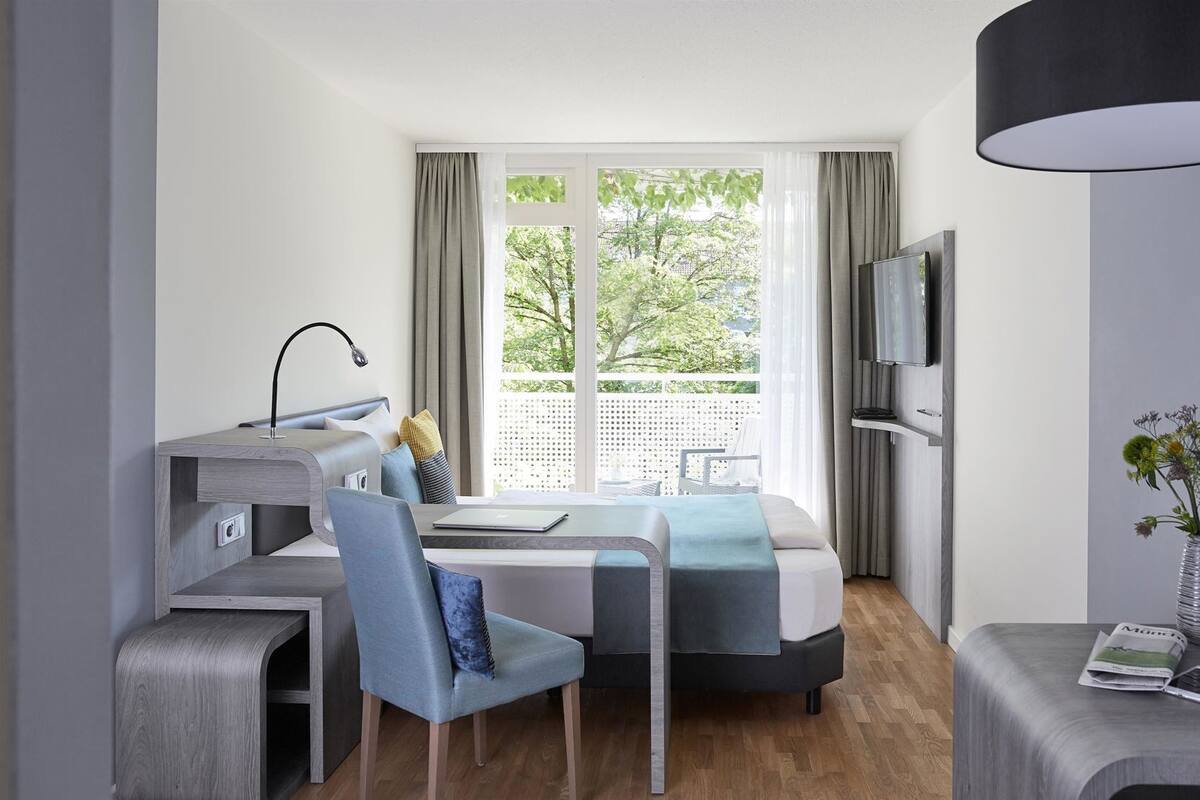 A modern hotel room with a bed, desk, chair, TV, and a small table with a vase.