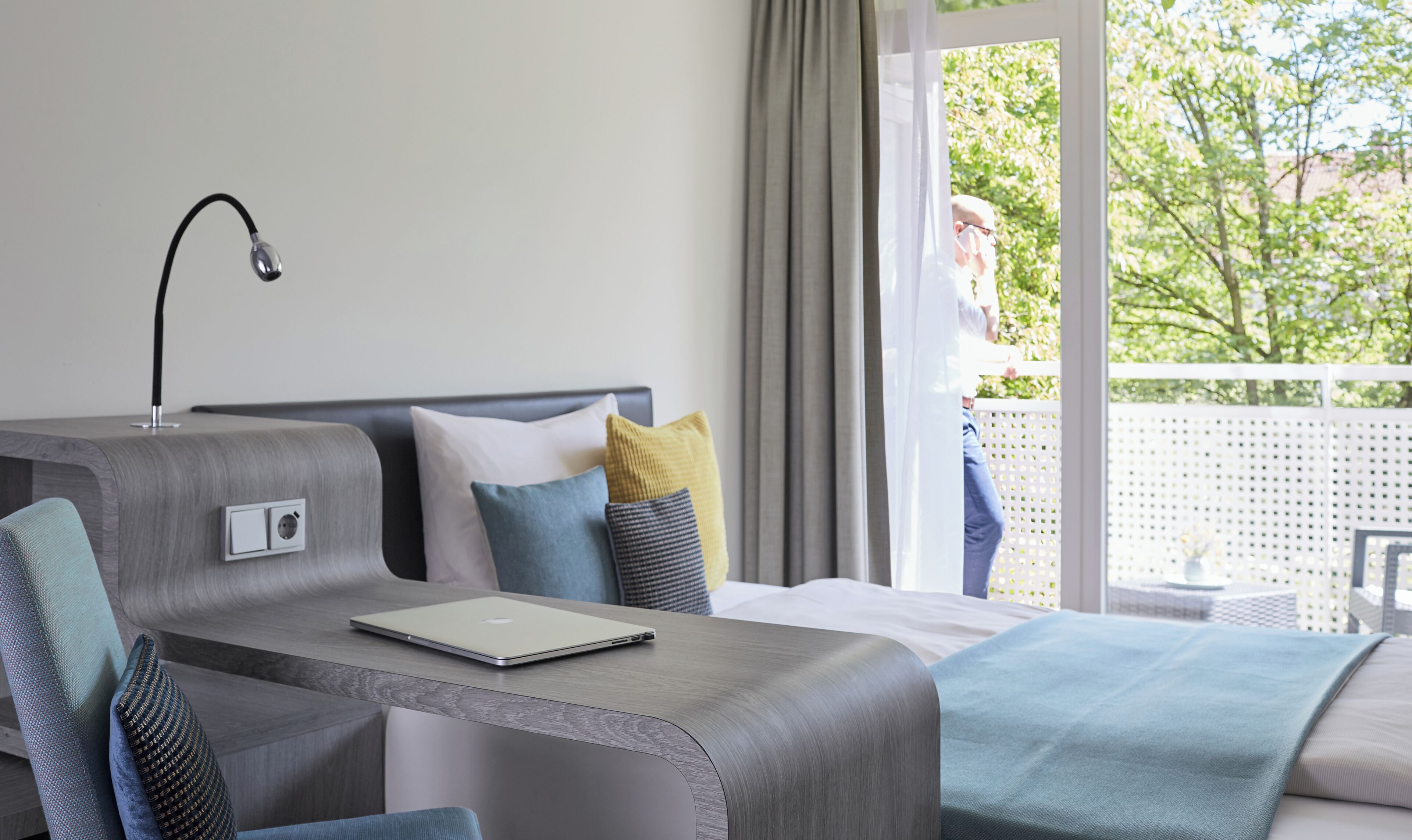 Une chambre d’hôtel moderne équipée d’un lit, d’un bureau avec un ordinateur portable, d’une chaise et d’une fenêtre donnant sur un paysage verdoyant.