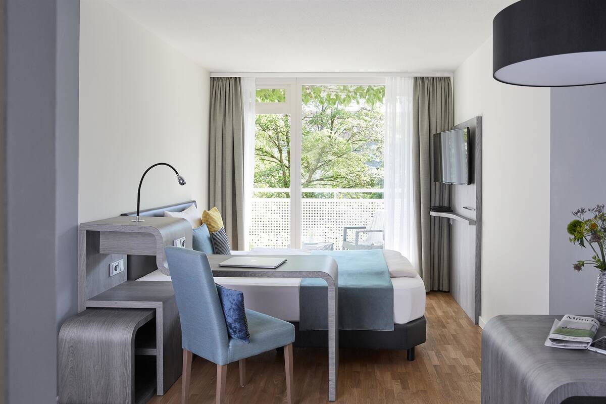 A modern hotel room with a bed, desk, chair, TV, and a vase with flowers.