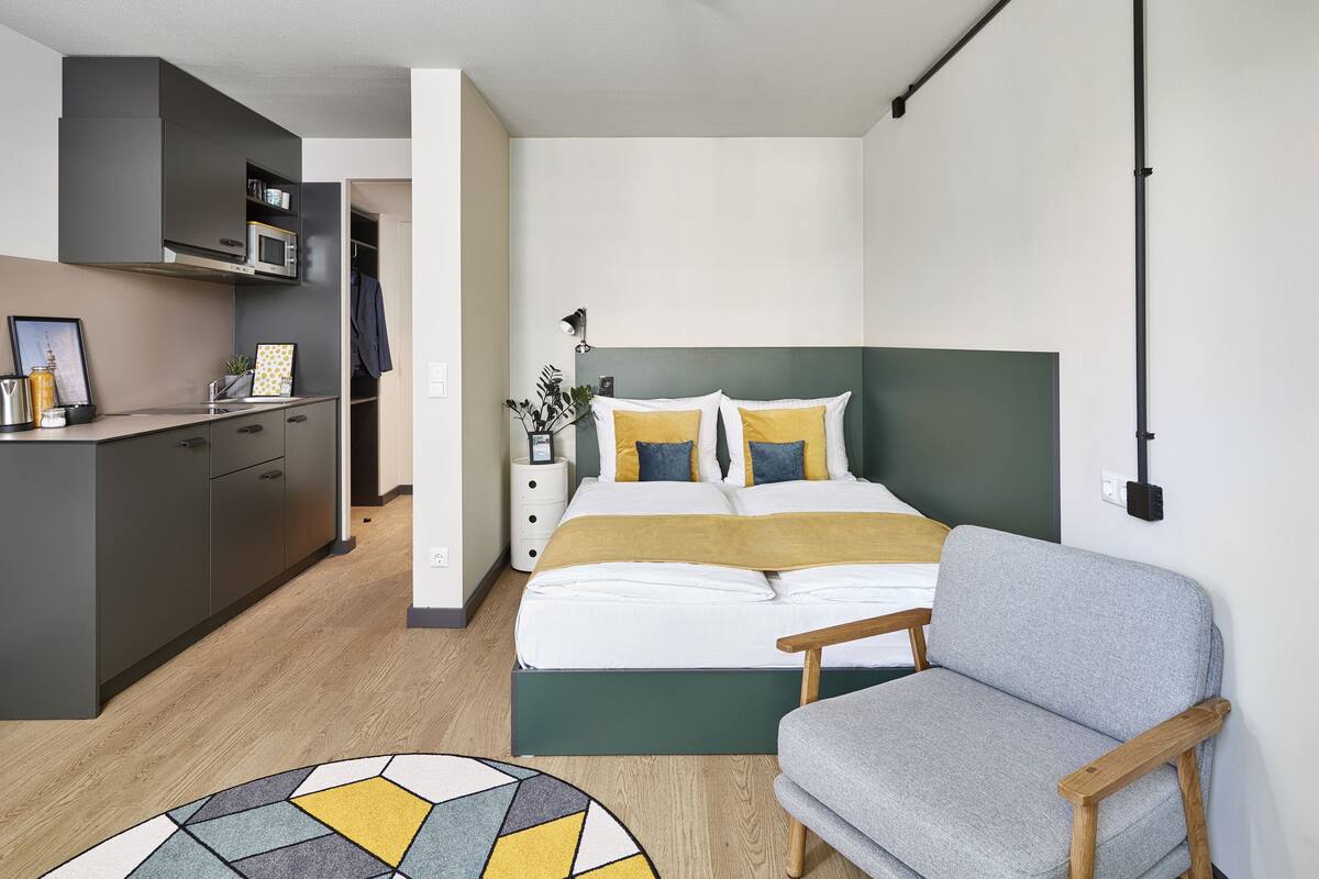 A compact hotel room with a kitchenette, a bed with a green headboard, a grey sofa, and a patterned rug.