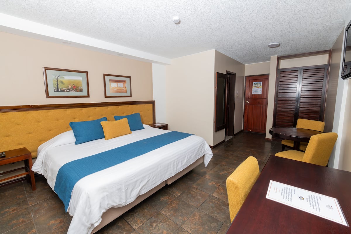 Habitación de hotel con cama, cabecera de madera, mesa de comedor con sillas y puerta que da a otra habitación.
