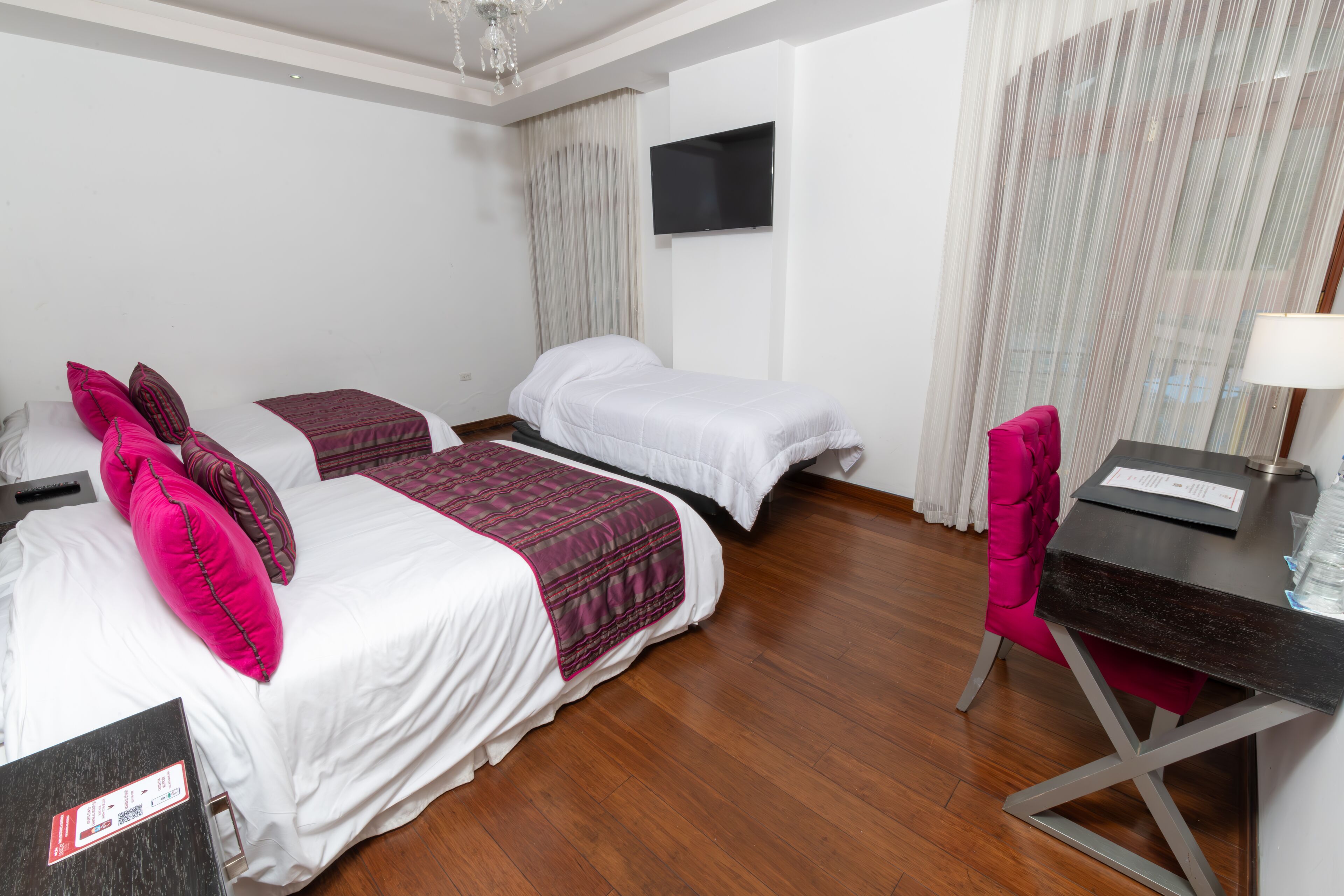 A hotel room with two beds, a desk, a chair, a TV, and a chandelier.