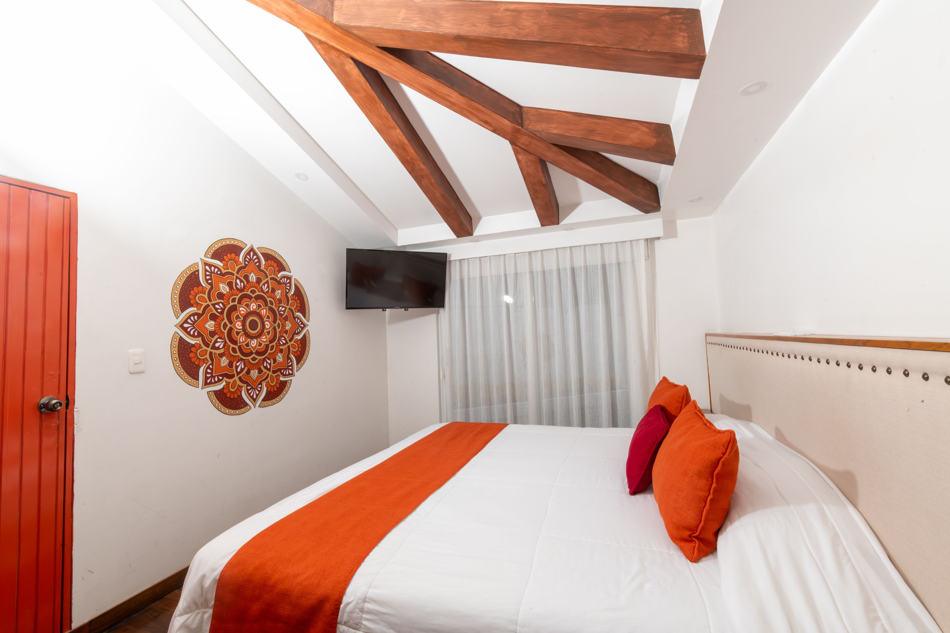 Un dormitorio con una cama, un televisor de pantalla plana, un elemento decorativo en la pared y estructura de vigas de madera en el techo.