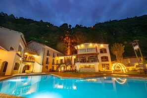 Et hotel med swimmingpool, flere balkoner og et bjerglandskab i baggrunden.