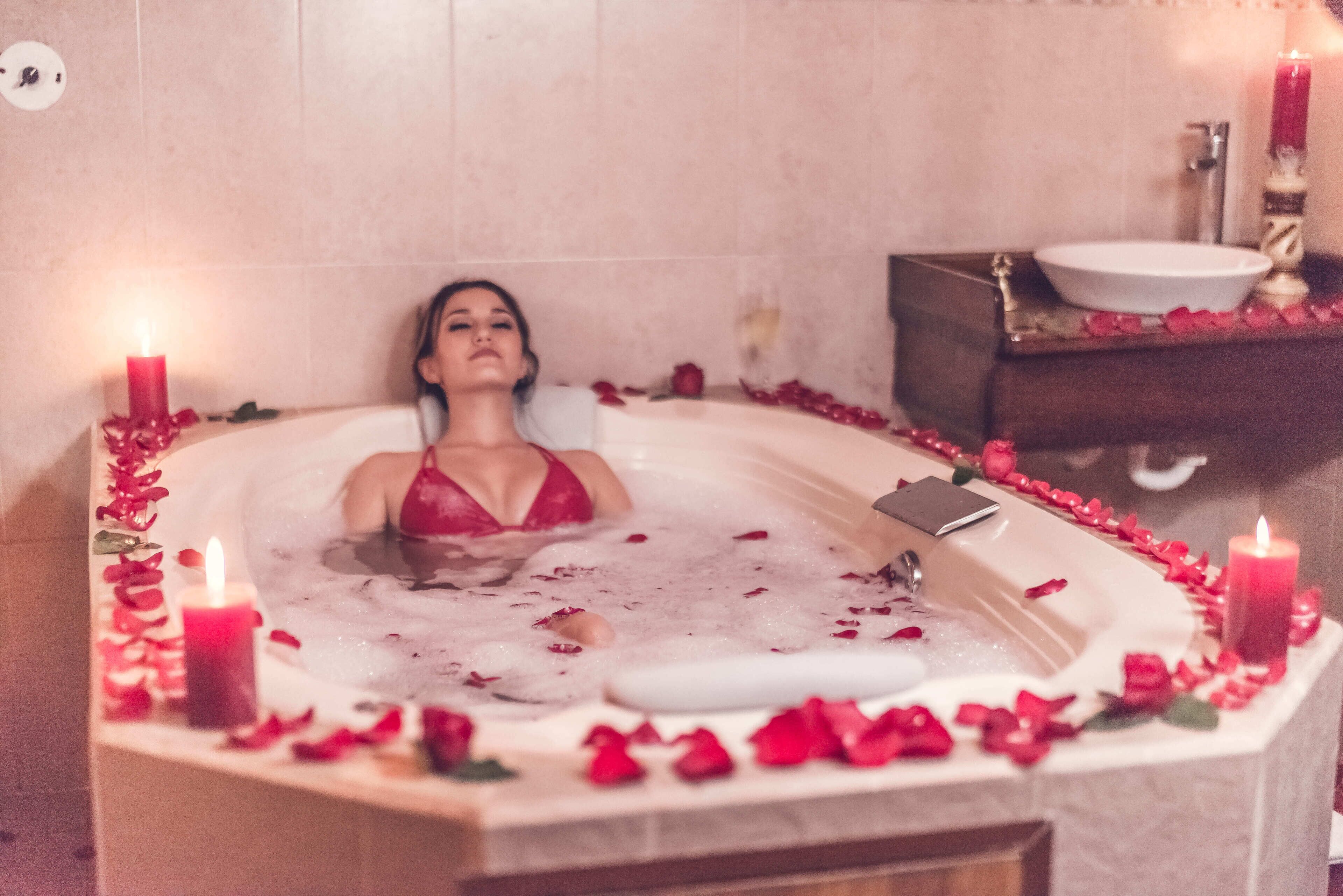 A woman relaxing in a bathtub filled with water and rose petals, surrounded by candles.