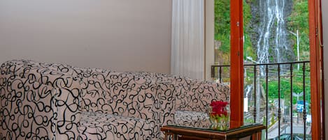Una sala de estar con un sofá estampado, una mesa de centro de vidrio y vistas a una cascada desde la puerta del balcón.