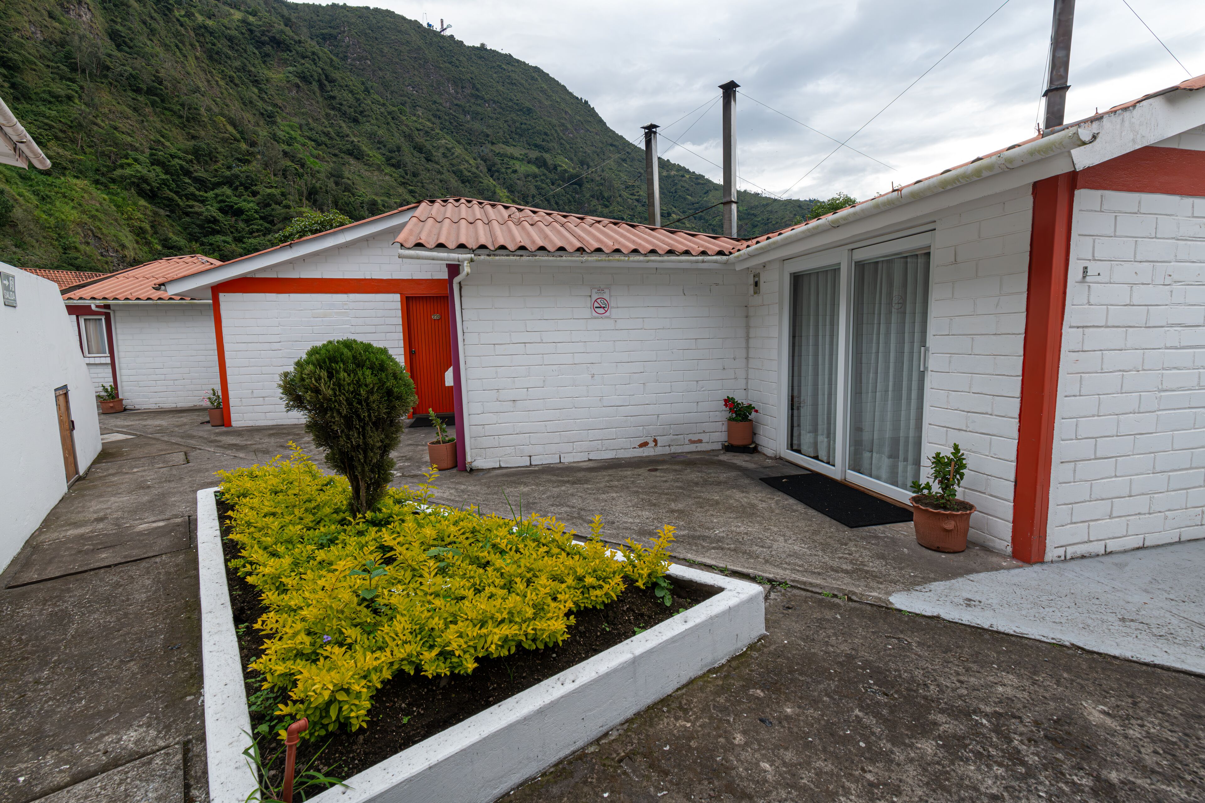 Edifici petit d'una sola planta amb una porta vermella i parets blanques, envoltat d'un jardí amb plantes grogues.
