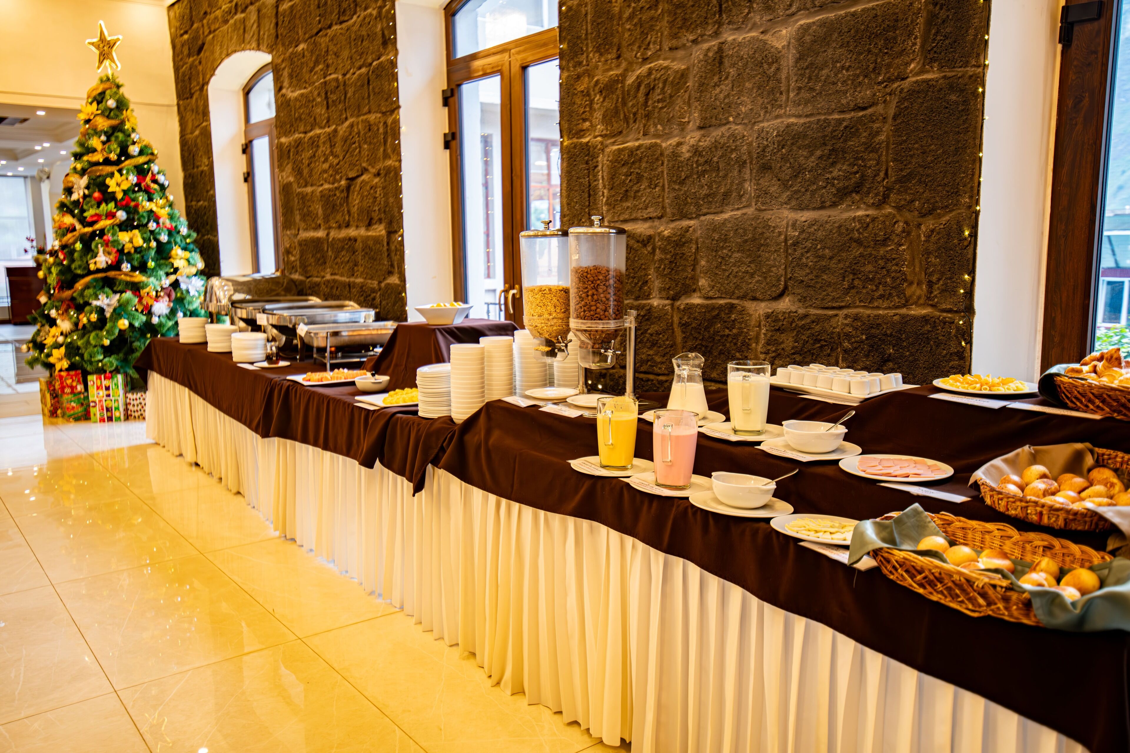 A buffet setup with a Christmas tree, food and drink dispensers, and baskets of bread.