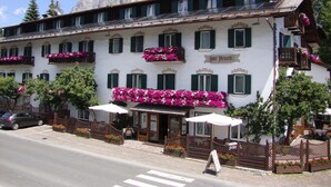 Ein Hotel mit heller Fassade, grünen Fensterläden und Balkonen, die mit rosa Blumen geschmückt sind.