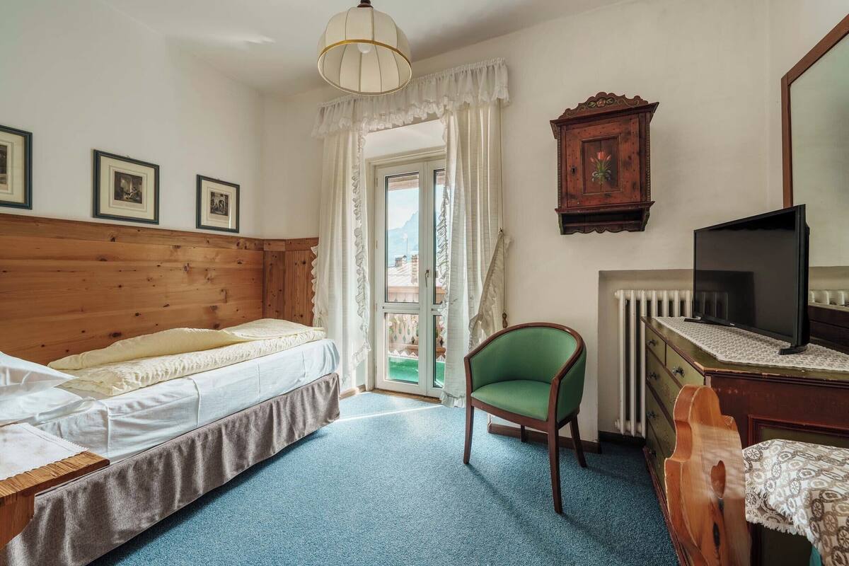 A bedroom with a wooden headboard, a bed with white linens, a green chair, a wooden dresser, a television, and a window with curtains.