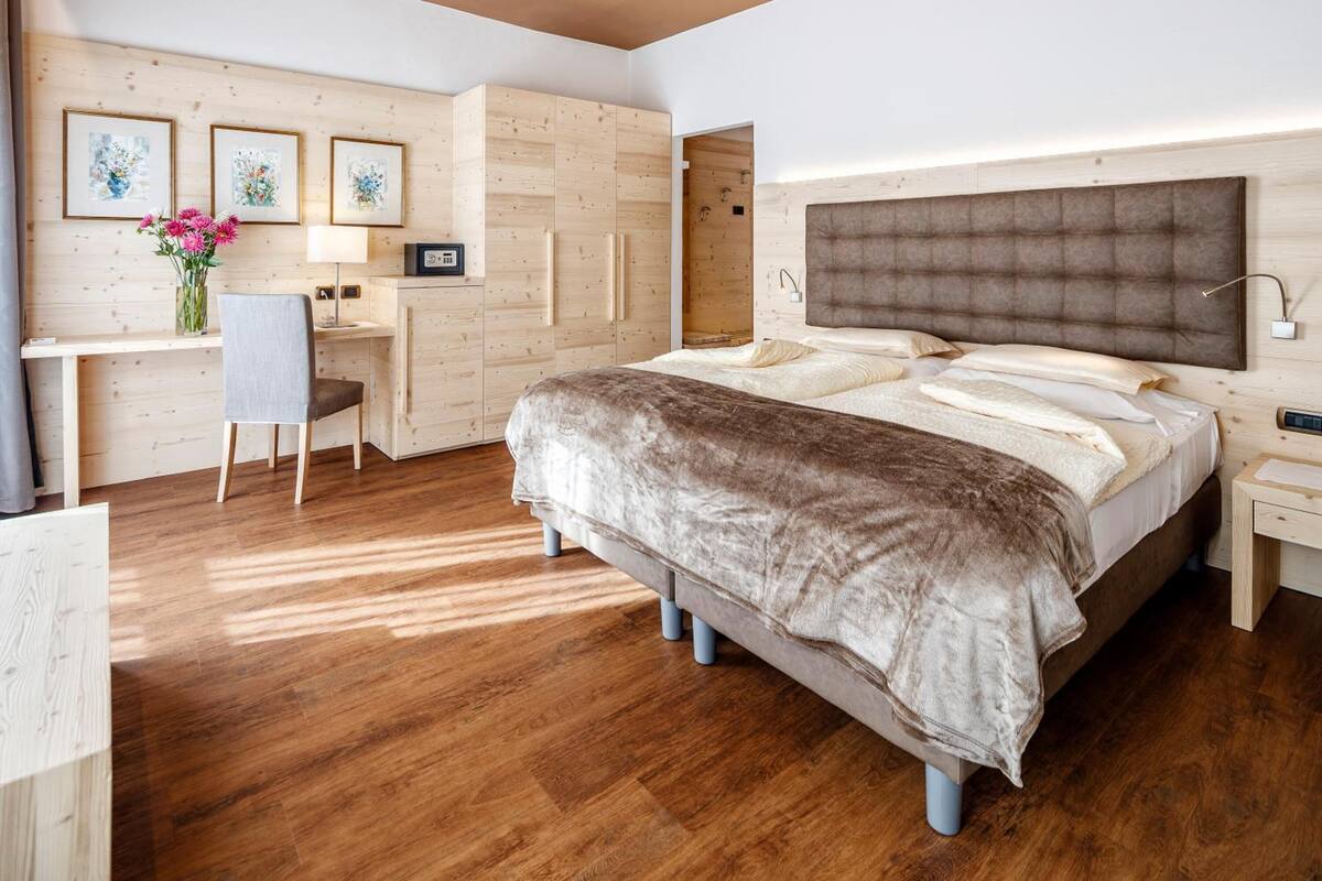 A bedroom with a large bed, a desk, and a chair. There are framed pictures on the wall and a vase with flowers.