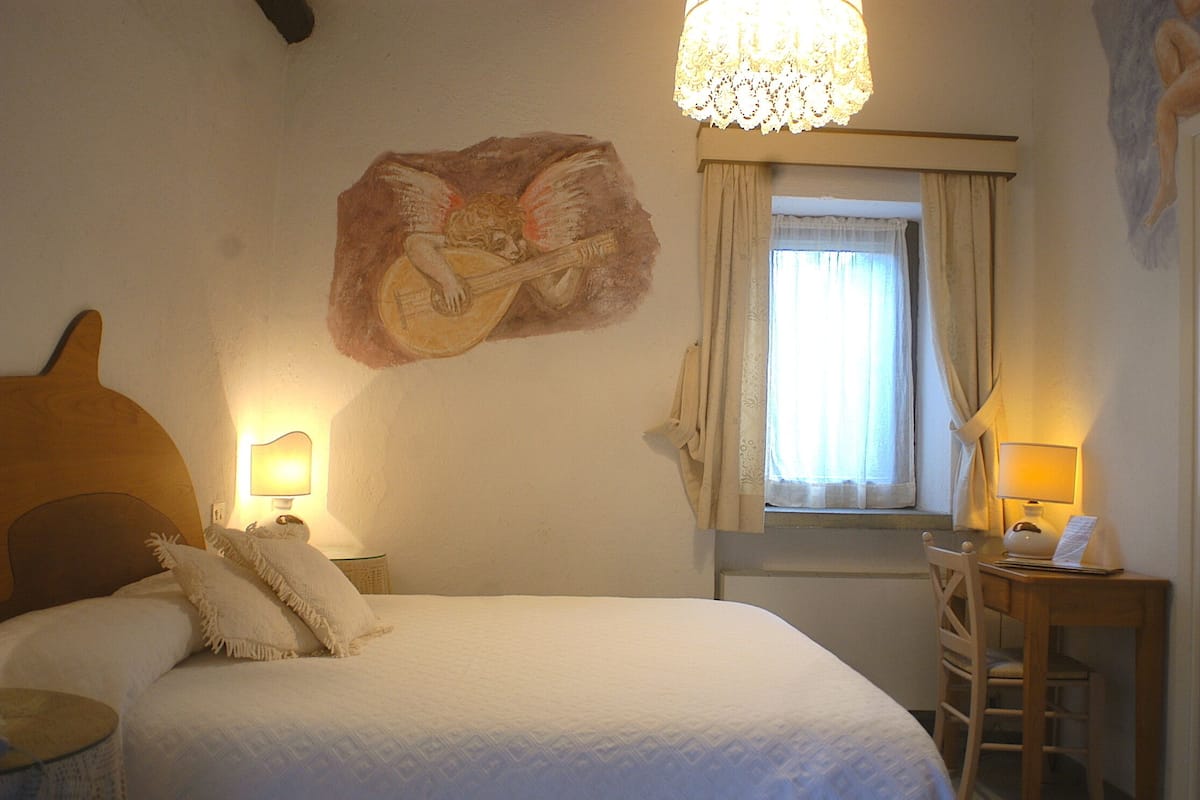 Een hotelkamer met een bed, twee bedlampen, een raam met gordijnen, een bureau met stoel en een muurschildering van een engel die gitaar speelt.