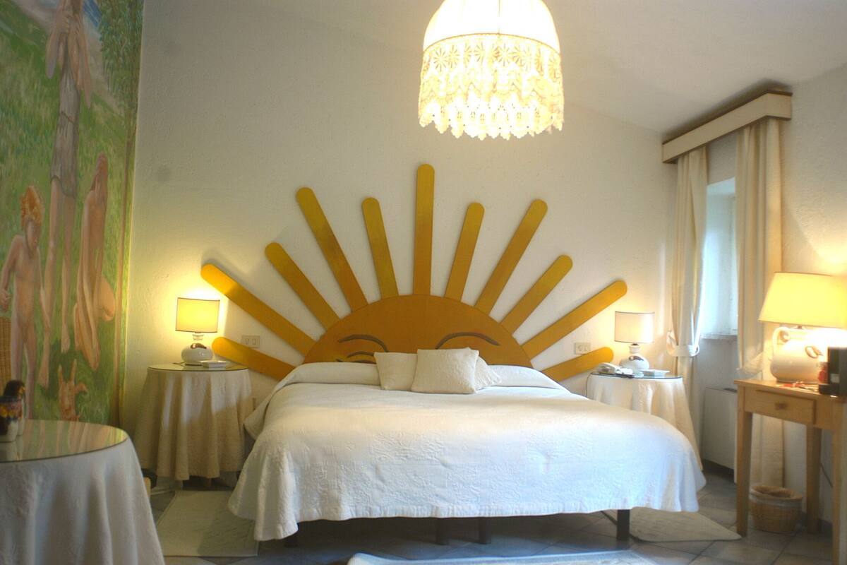 Hotelkamer met een zonneschijnbord boven het bed, twee nachtkastjes met lampen, een groot schilderij aan de linker muur en een kroonluchter.