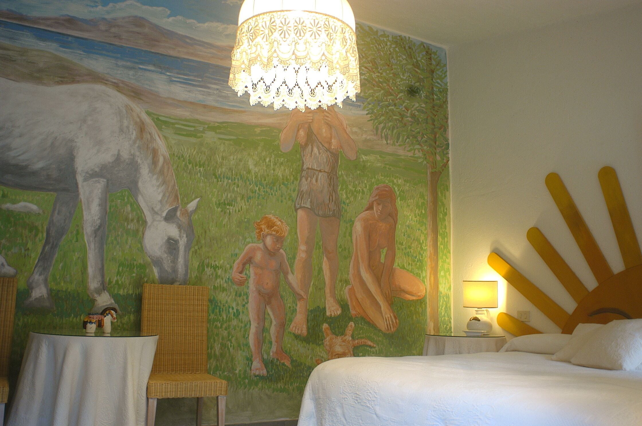 A hotel room with a mural, a bed, a nightstand, a lamp, and a wicker chair.