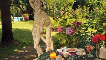 A garden setting with a statue, a table with breakfast items, and lush greenery.