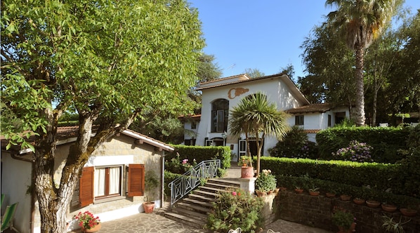 A traditional Mediterranean-style villa with a stone facade, a central staircase leading to the entrance, and a well-maintained garden with various plants and flowers.