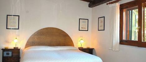 Quarto com cama grande, cabeceira de madeira, luminárias de cabeceira, quadros nas paredes e uma janela com cortinas.