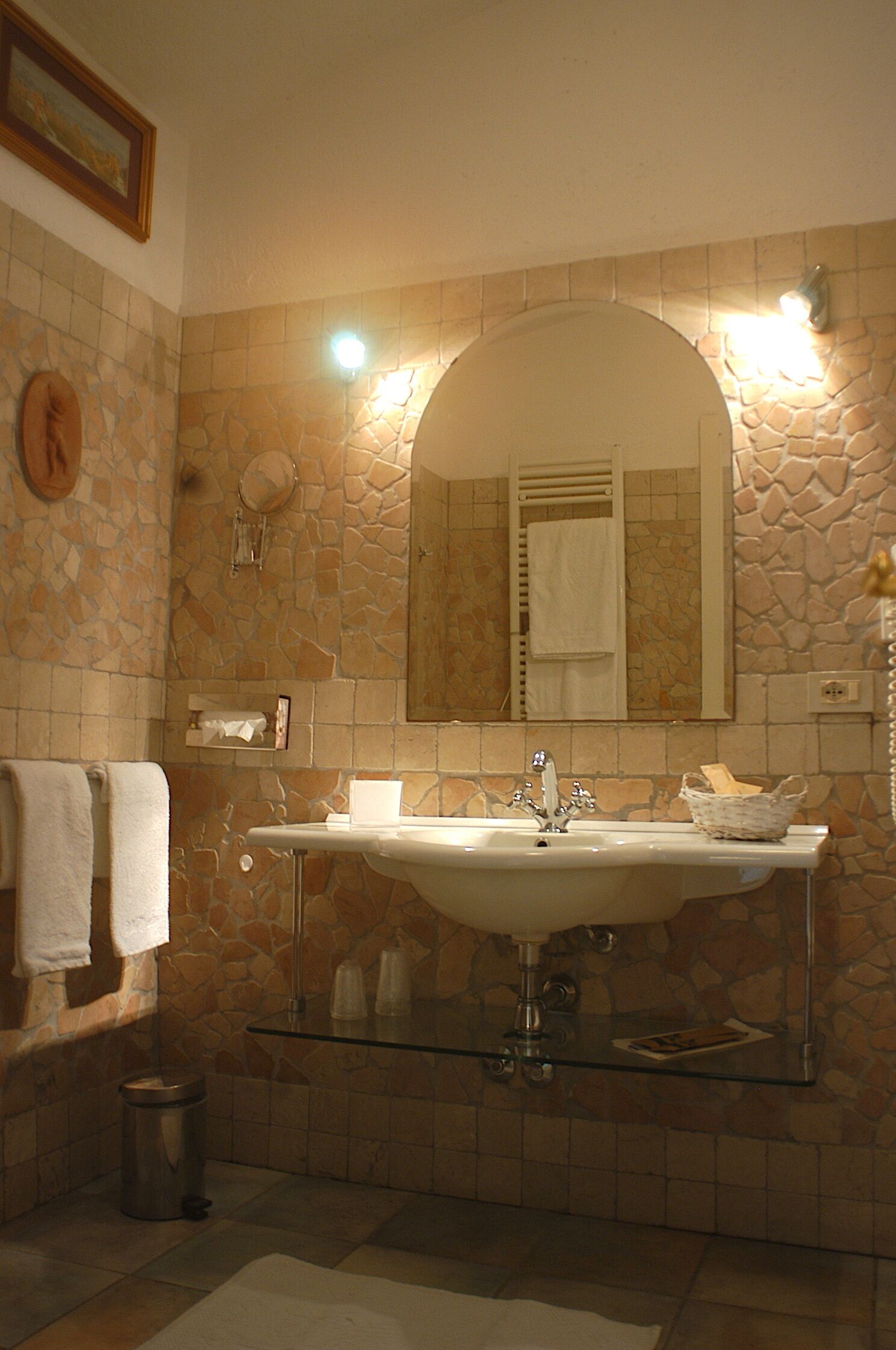 Un baño con pared de piedra, un espejo grande, lavamanos doble y un cubo de basura.