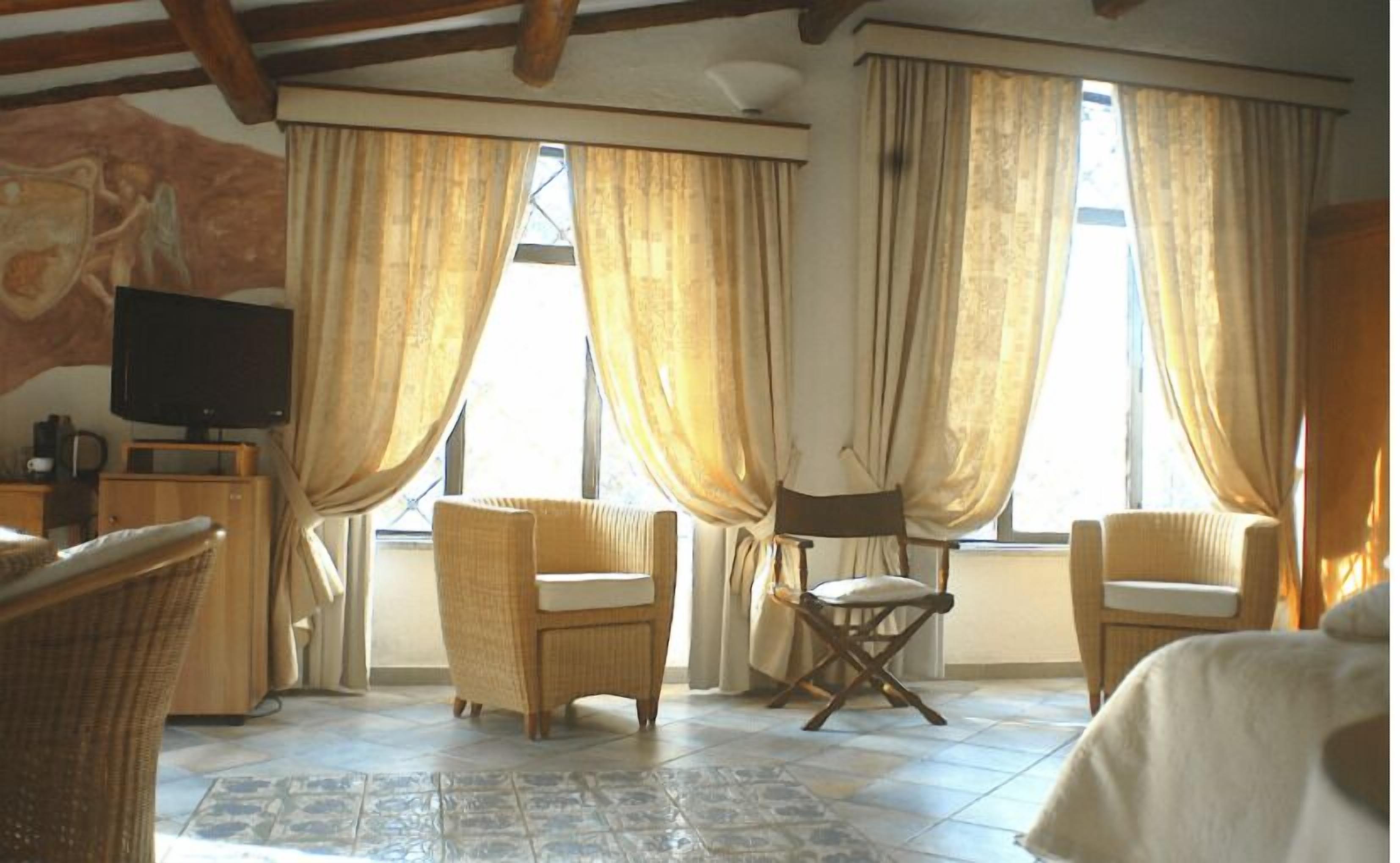 Una habitación con televisión, sillas de mimbre, una mesita y ventanas amplias con cortinas.