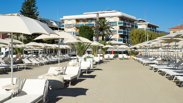 A beachside hotel with rows of sun loungers and umbrellas, a palm tree, and a building with balconies in the background. - Hotel Garden Lido (Loano)