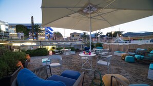 A beachside outdoor seating area with wicker furniture, umbrellas, and a view of the sea and buildings in the background. - Hotel Garden Lido (Loano)