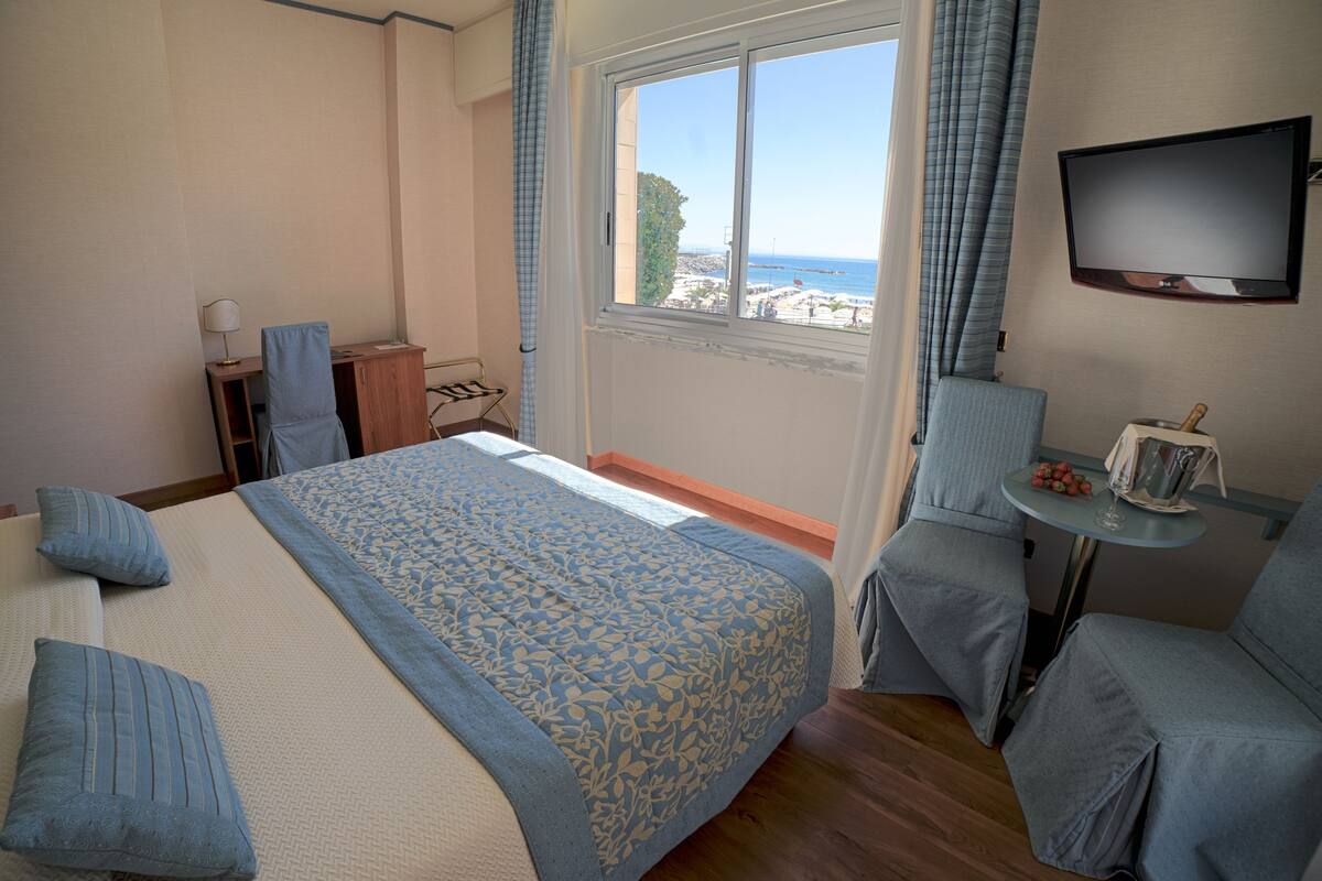 A hotel room with a bed, a desk, a chair, a TV, and a view of the beach.