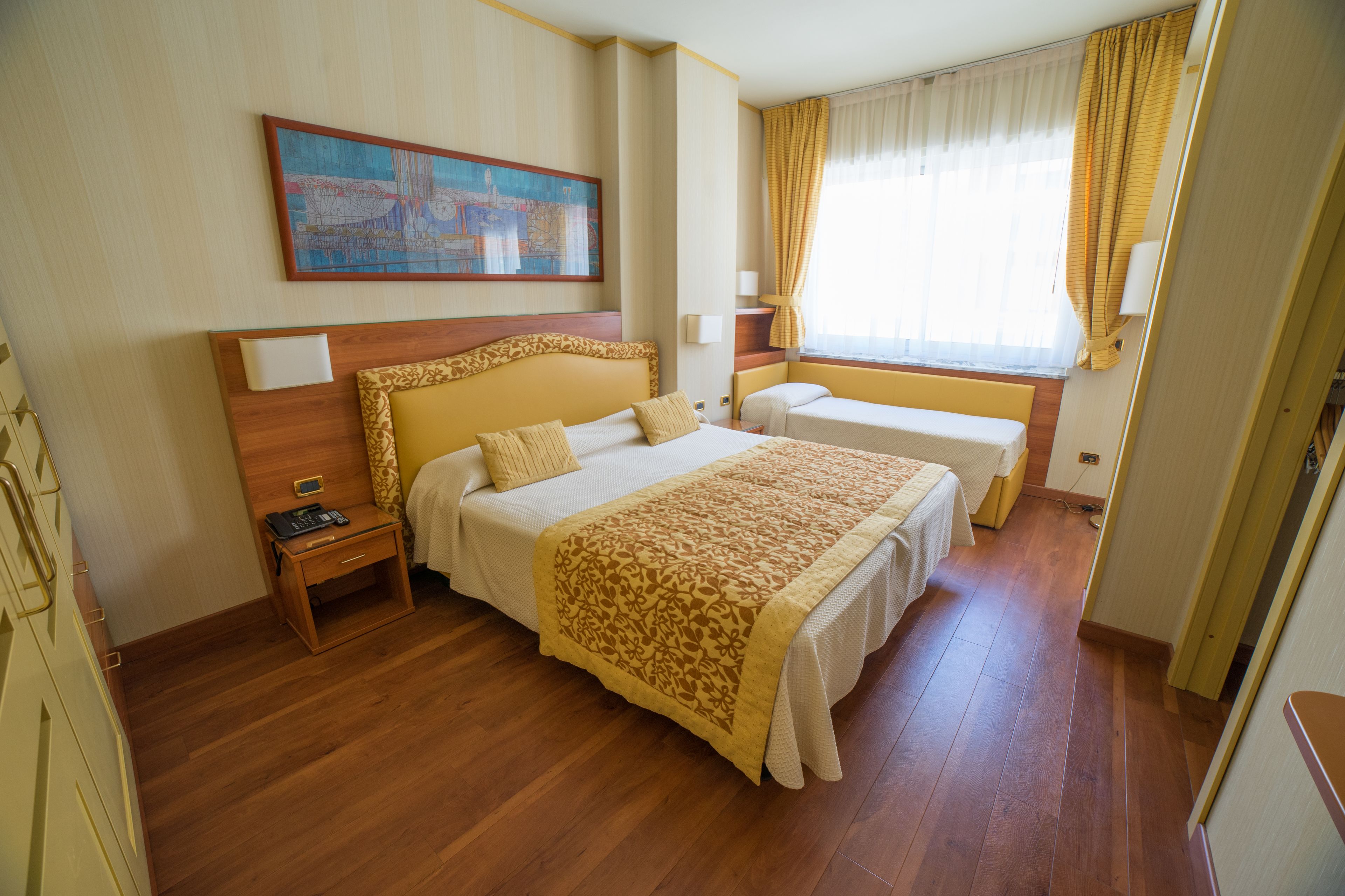 Habitación de hotel con una cama grande, una cama más pequeña, una mesita de noche, un cuadro en la pared y una ventana con cortinas.