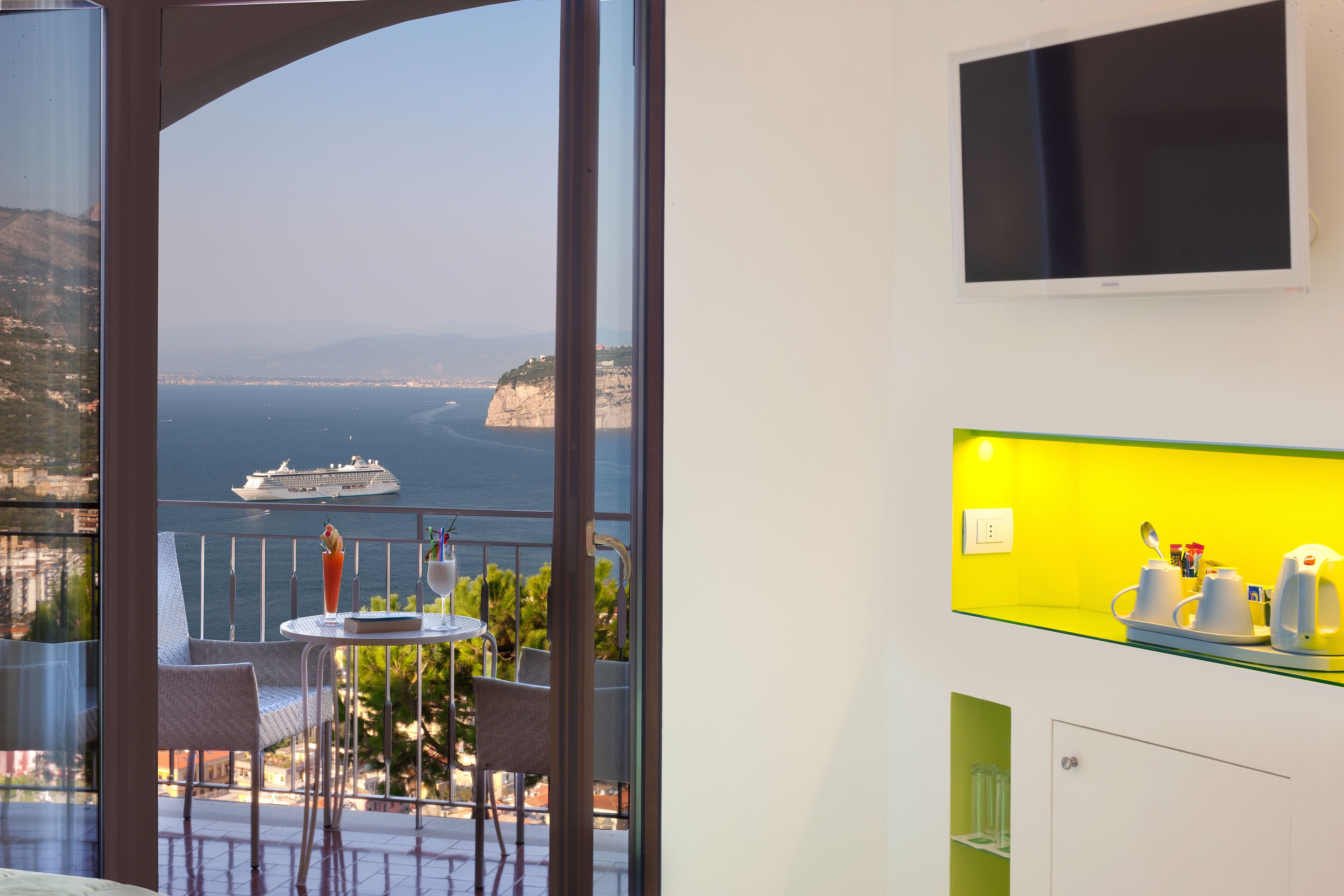 Un balcon avec vue sur la mer, un navire et le paysage urbain. On y trouve une table avec des chaises et un verre contenant une boisson.