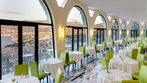 Un restaurante con mesas redondas y sillas verdes, amplios ventanales de arco y vistas a la ciudad.
