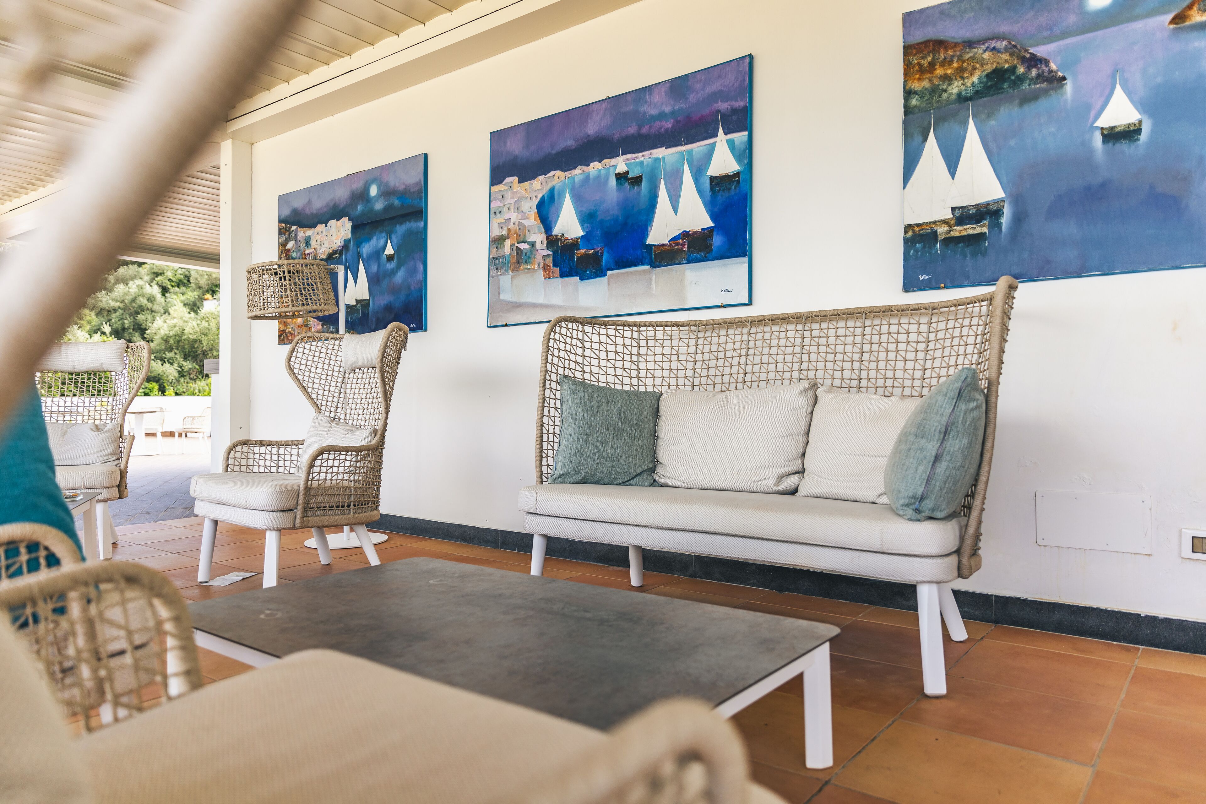A cozy lounge area with wicker furniture, a sofa, and a coffee table. There are three paintings on the wall depicting sailboats.