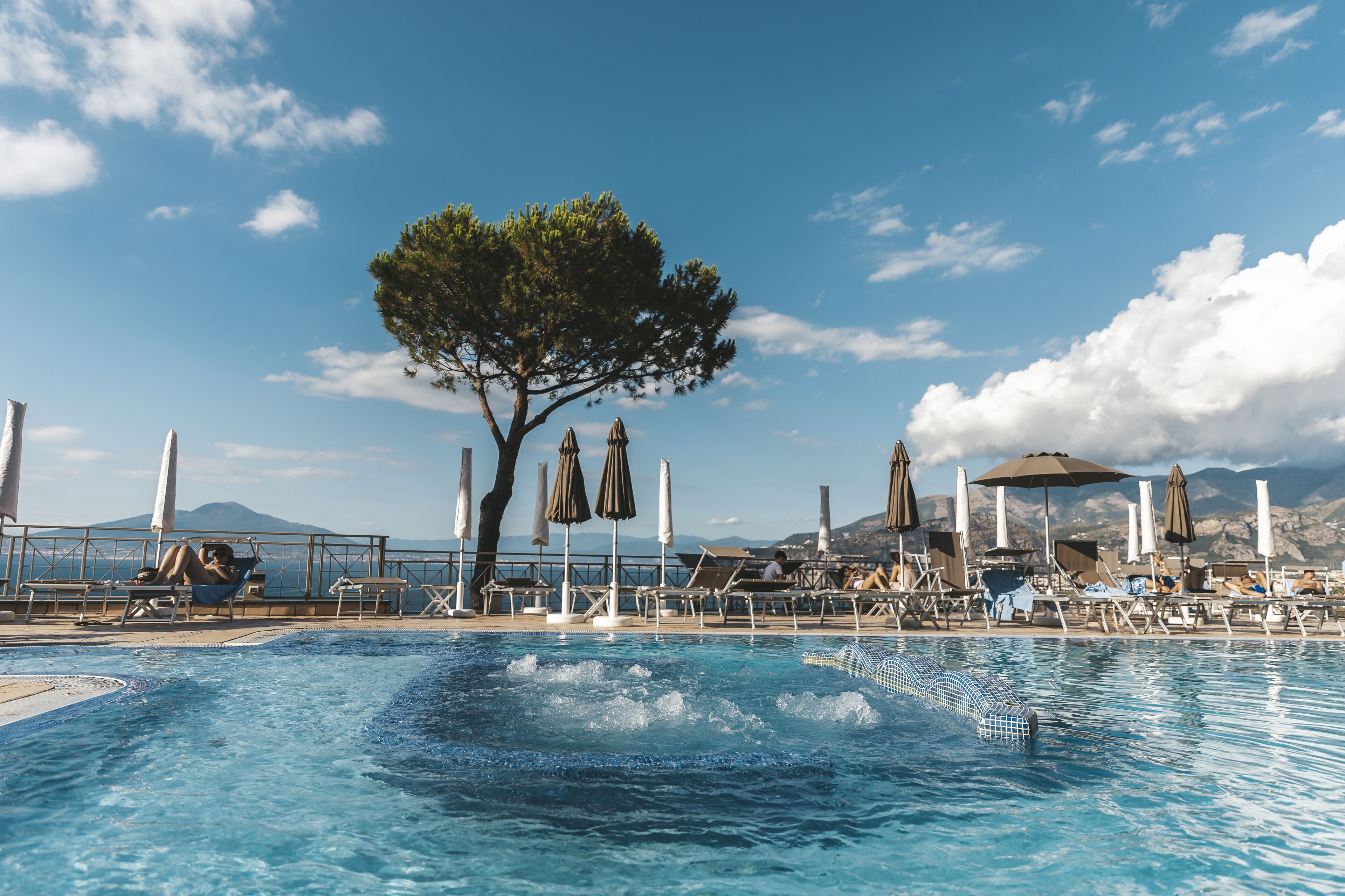 A poolside area with lounge chairs and umbrellas, a large tree, and a mountainous backdrop.