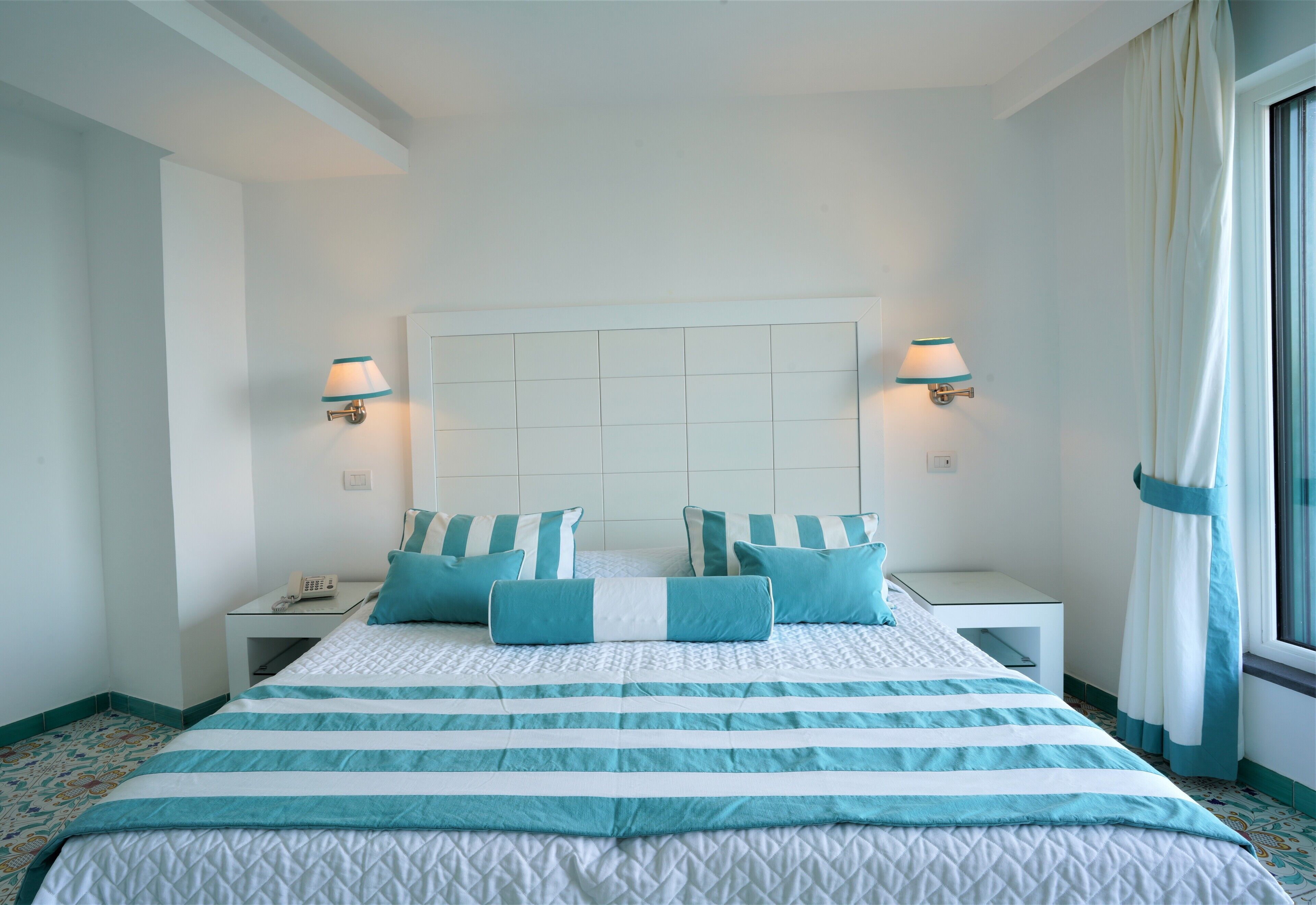 A hotel room with a large bed, two bedside tables, a nightstand, a window with curtains, and wall-mounted lamps.