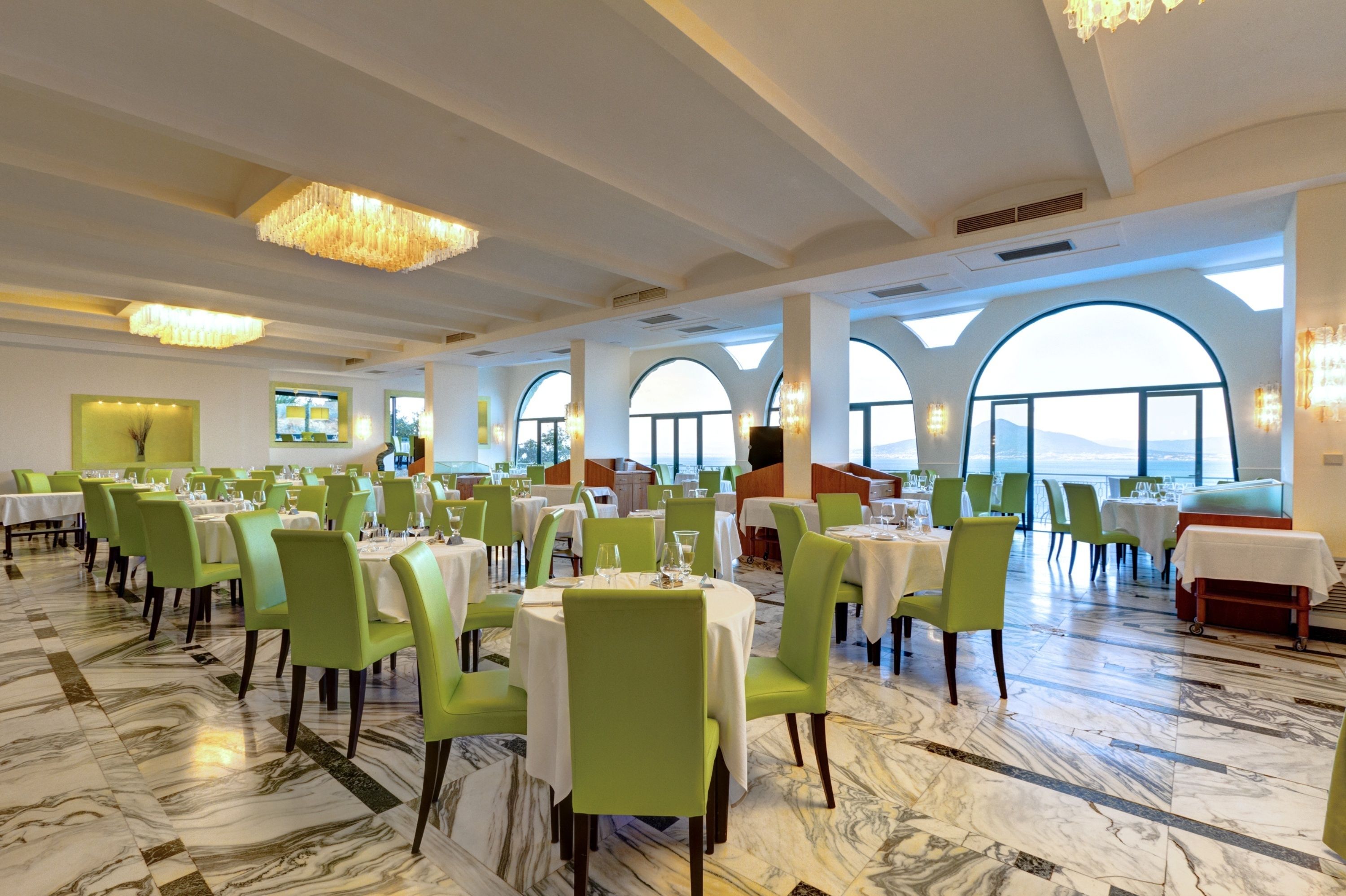 A spacious dining area with green chairs and white tables, large arched windows, and a chandelier.