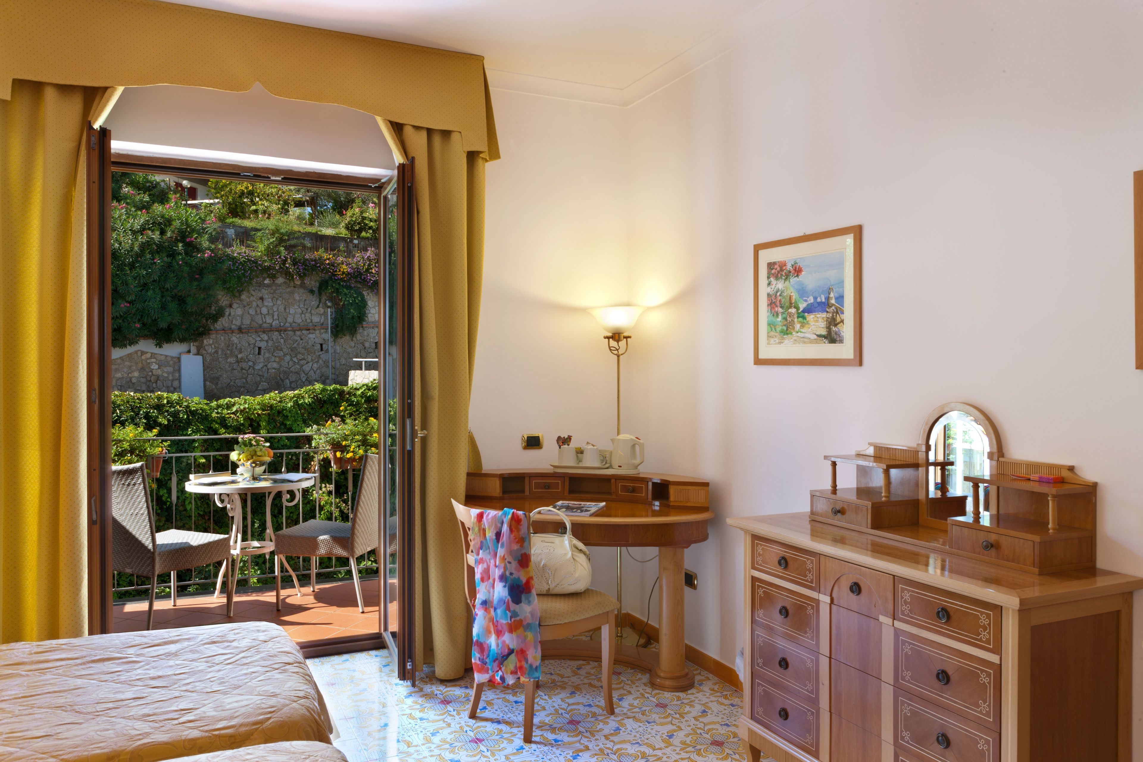 A hotel room with a bed, a desk, a chair, a dresser, and a balcony with a table and chairs.