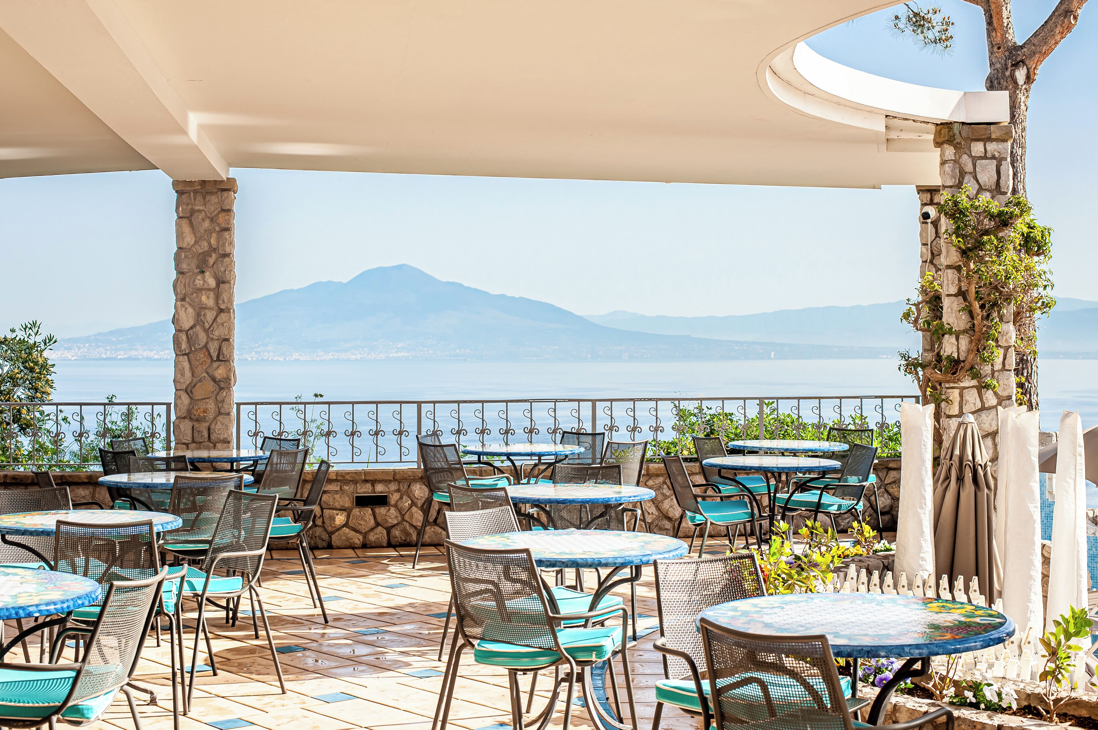 Une terrasse extérieure avec des tables et des chaises rondes, un pilier en pierre et une vue sur la mer et les montagnes.