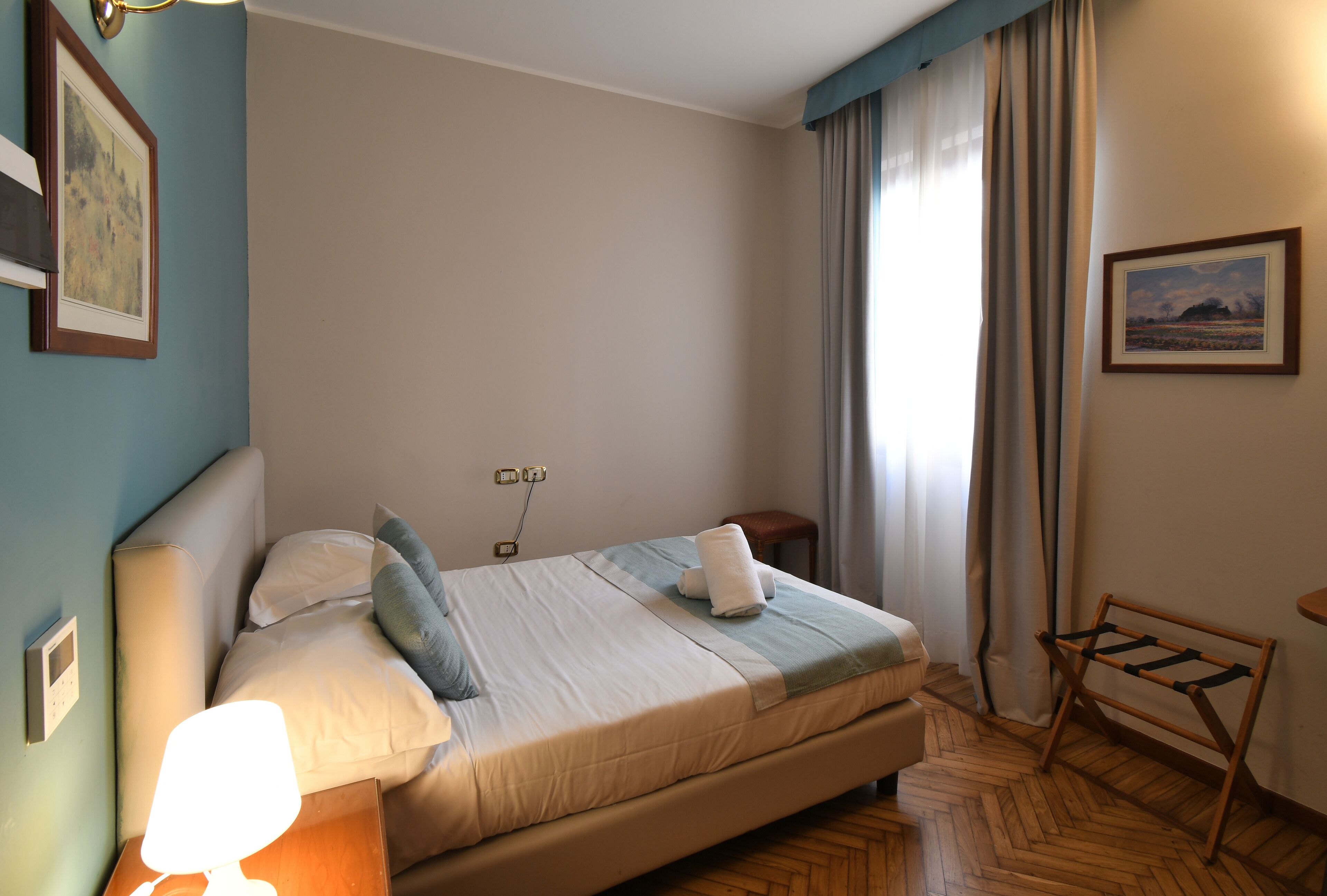 Cama bem arrumada com travesseiros e manta, mesa de cabeceira com abajur, piso de madeira e janela com cortinas.
