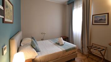 Cama bem arrumada com travesseiros e manta, mesa de cabeceira com abajur, piso de madeira e janela com cortinas.