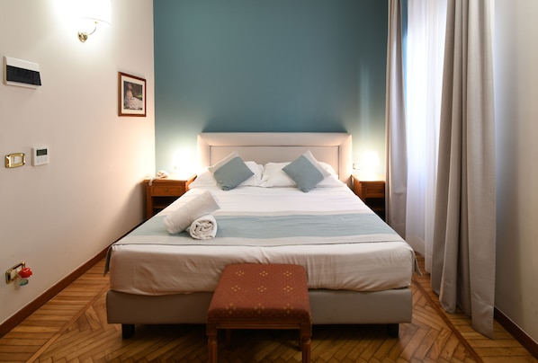 A neatly made bed with pillows, a bedside table, a wall-mounted light, and a wooden floor.