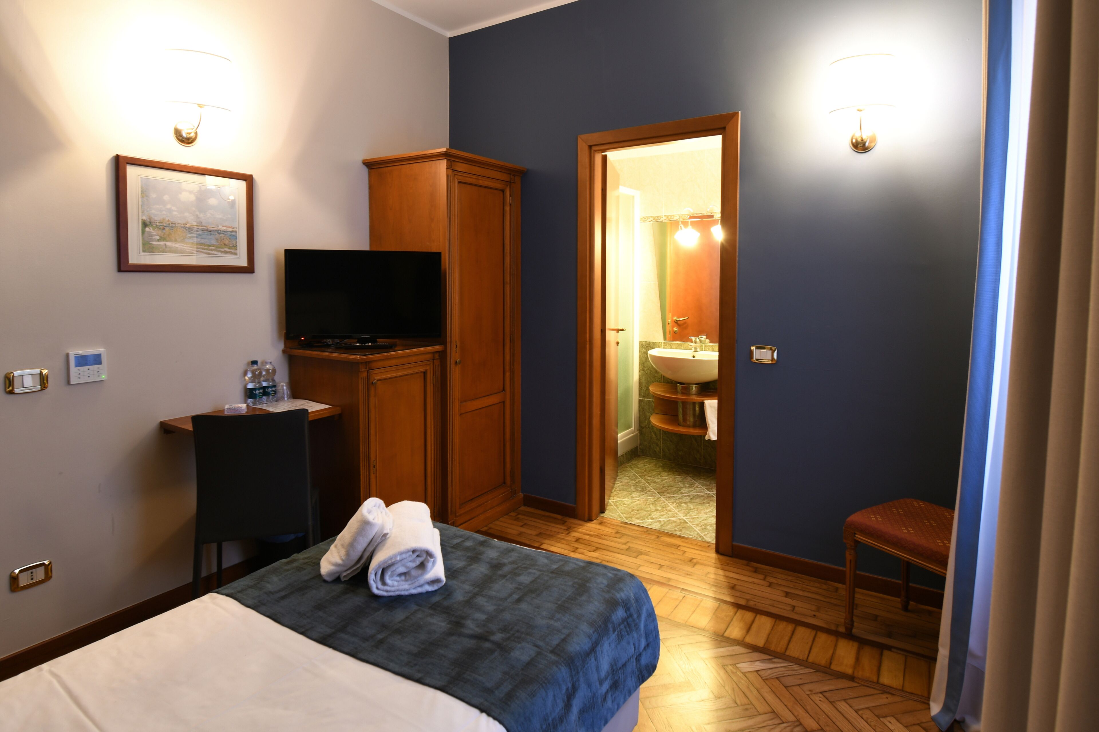 A hotel room with a bed, a television, a desk with a chair, a wardrobe, and a bathroom visible through an open door.