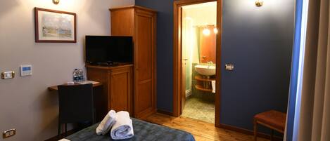 A hotel room with a bed, a television, a desk with a chair, a wardrobe, and a bathroom visible through an open door.