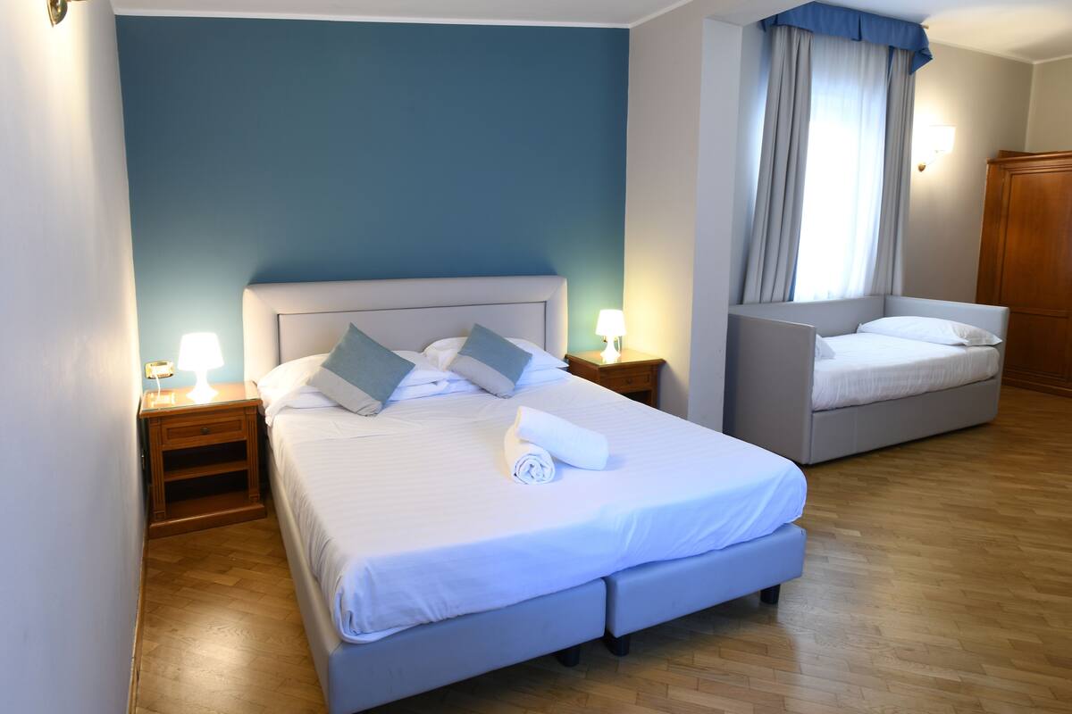 A double bed with a headboard, two bedside tables with lamps, a small bedside table with a lamp, a wooden wardrobe, and a window with curtains.