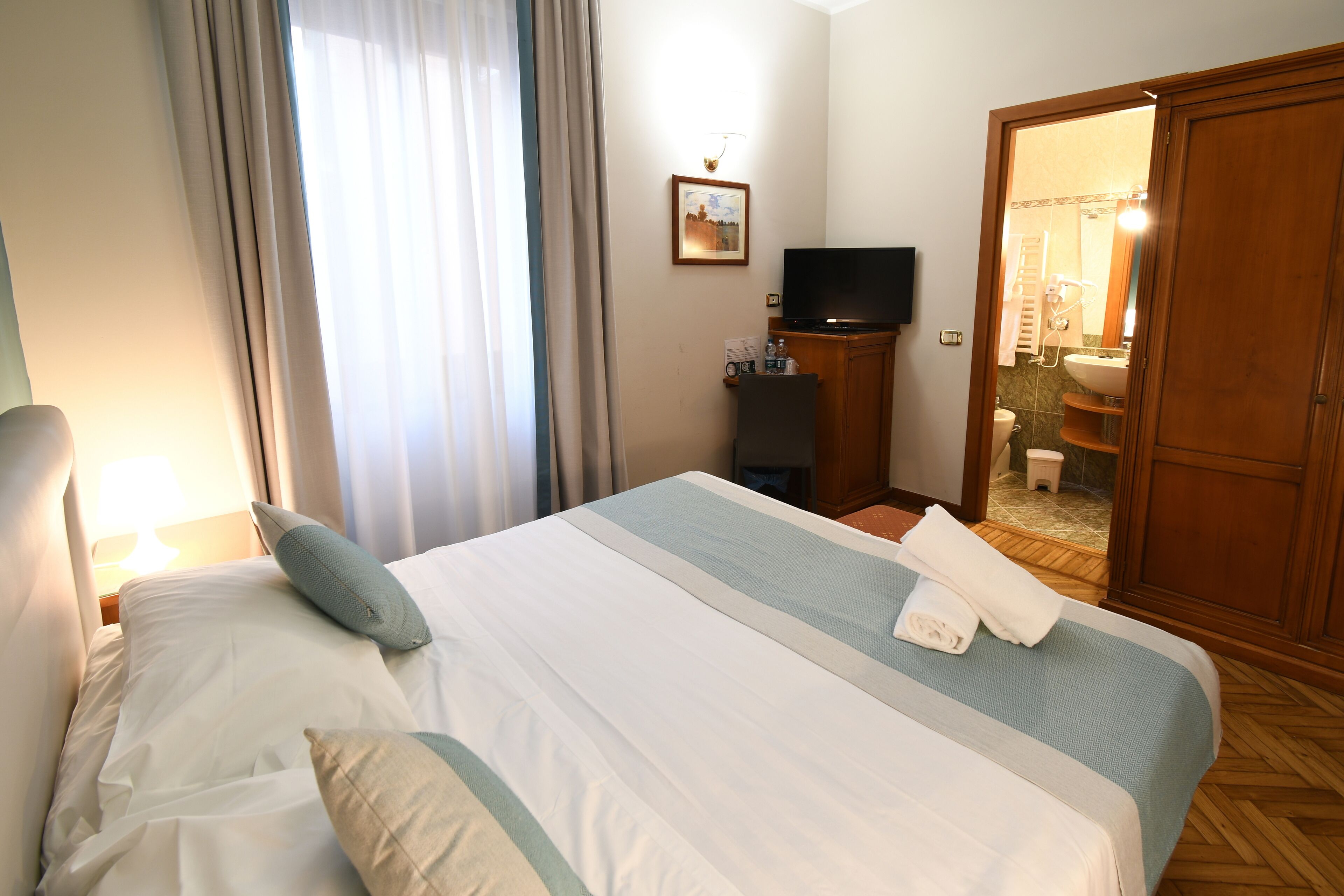 Camera d'albergo con un letto, una televisione, un tavolino con una lampada e un bagno visibile tramite una porta aperta.