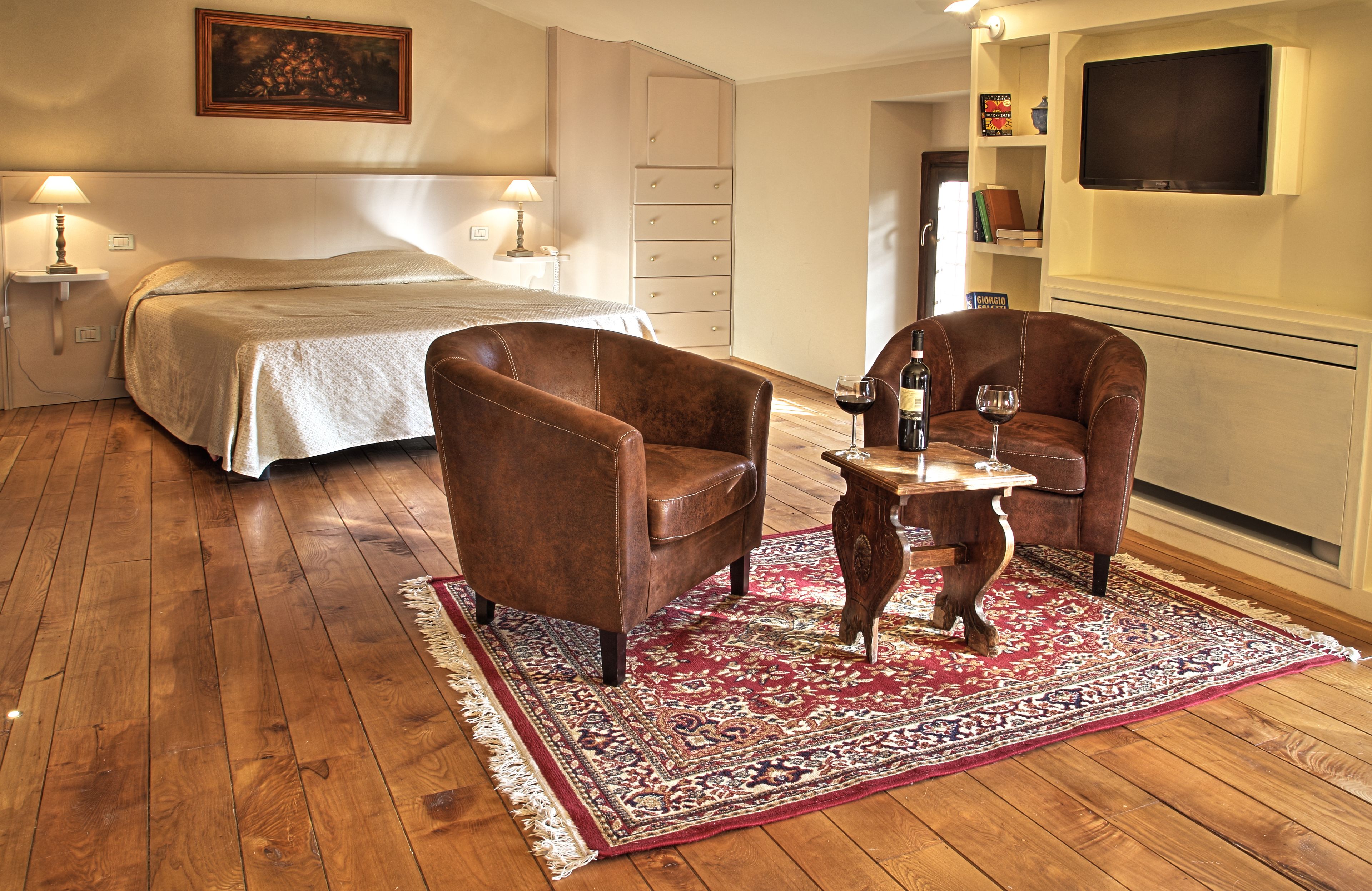 A hotel room with a bed, two leather chairs, a small table with wine, a TV, and a bookshelf.