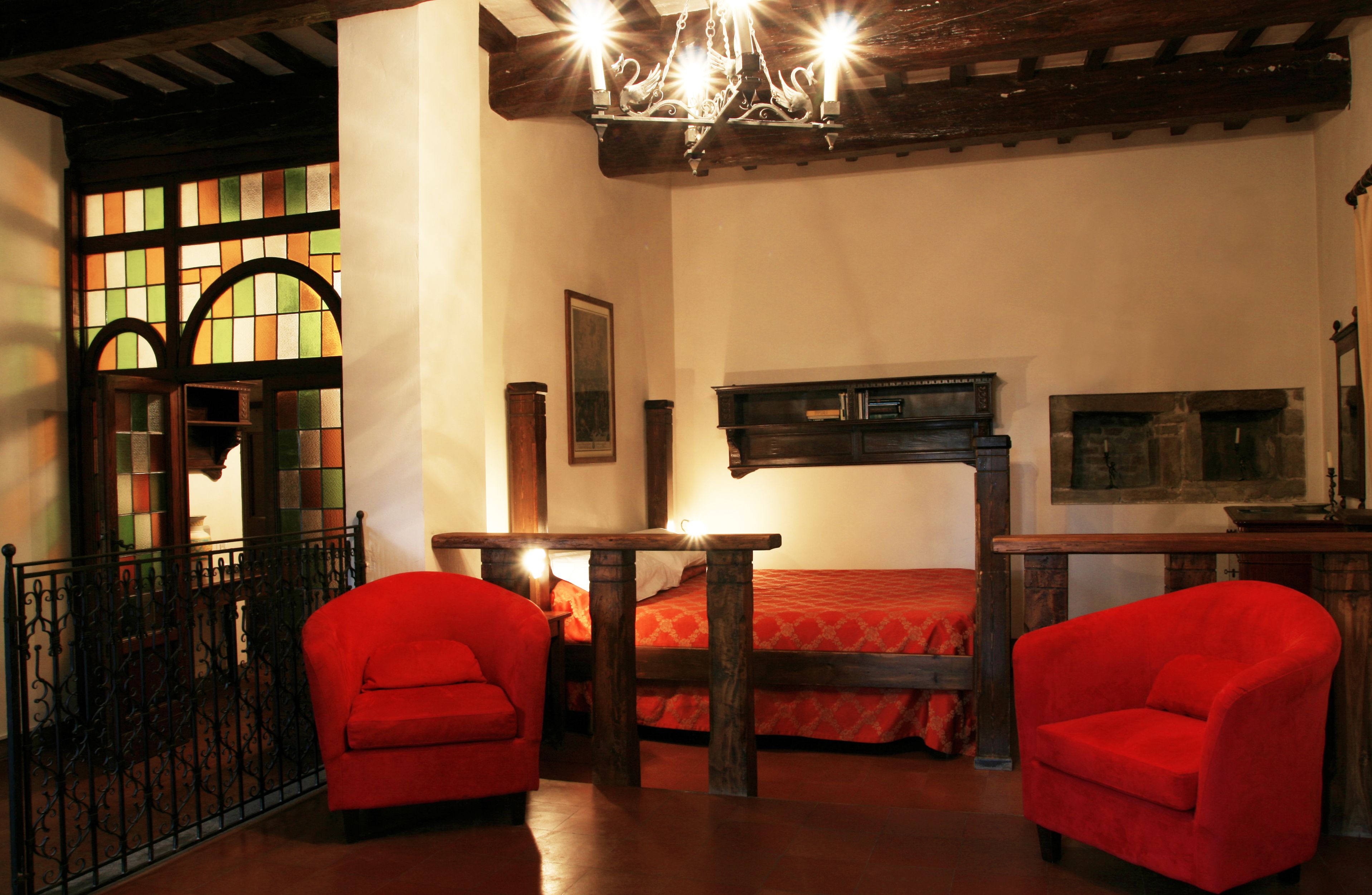 Una habitación con una cama, dos sillones rojos, una mesa de madera y una araña.