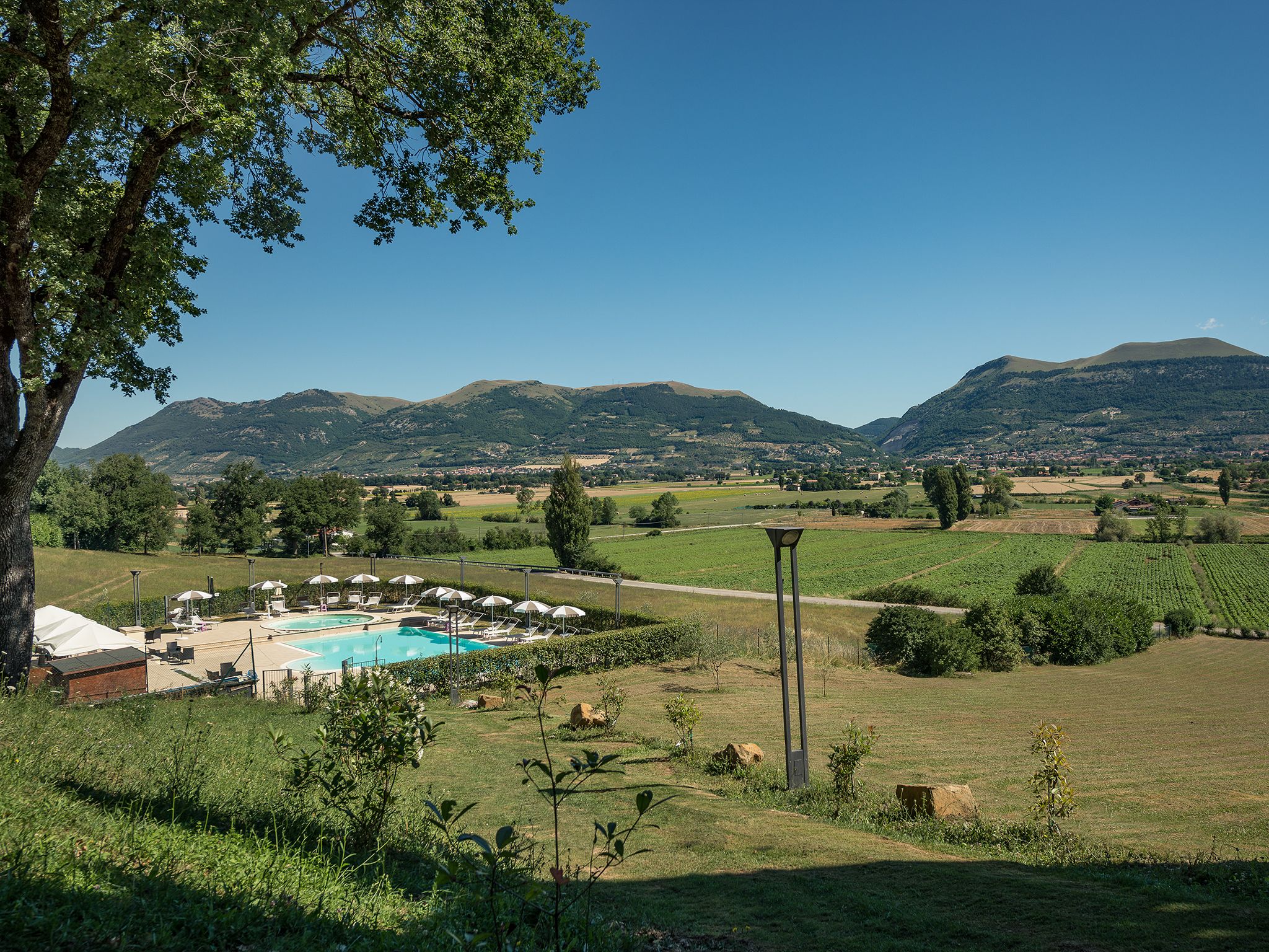 A pool area with lounge chairs and umbrellas, surrounded by green fields and mountains in the background.