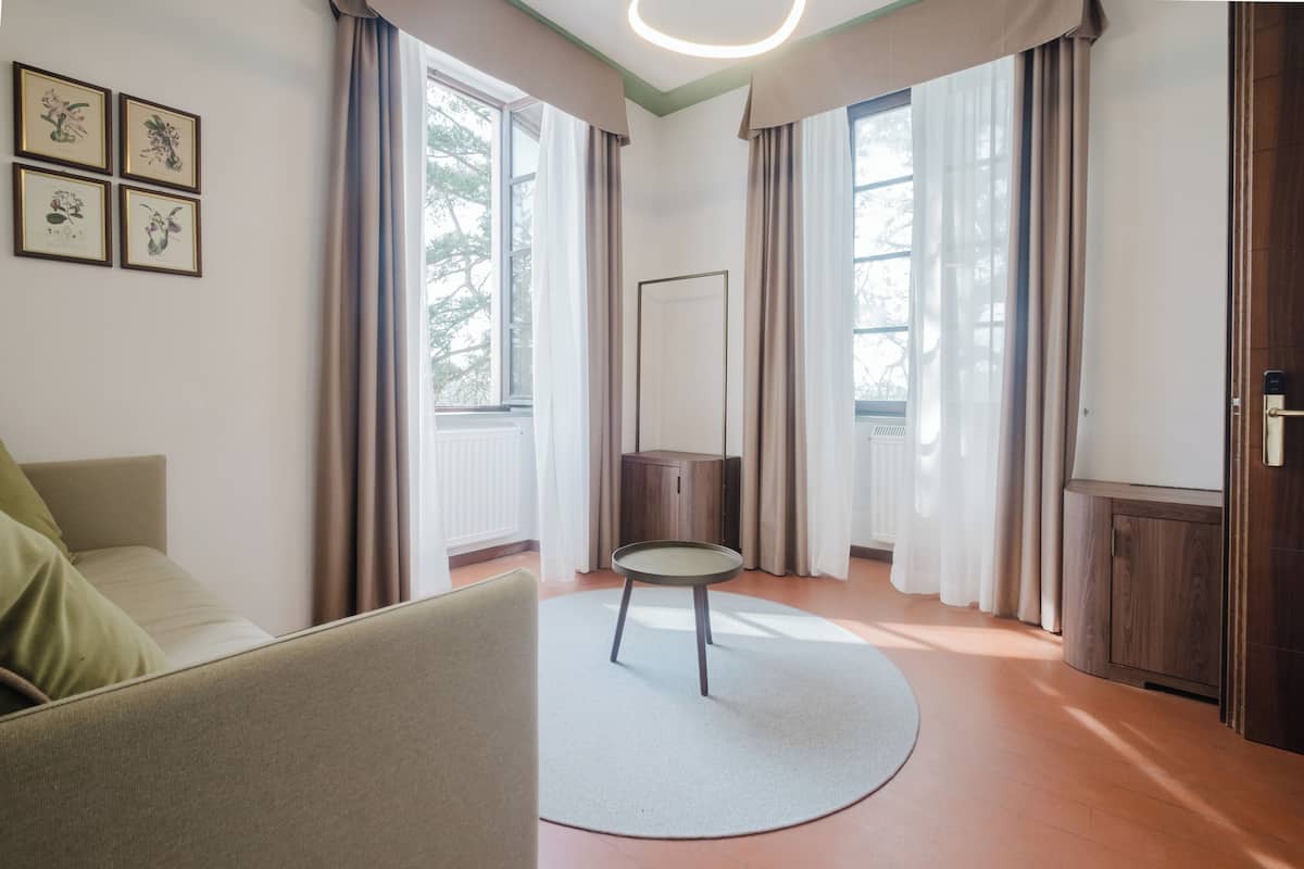 A modern hotel room with a sofa, a small round table, a cabinet, and a large window with curtains.