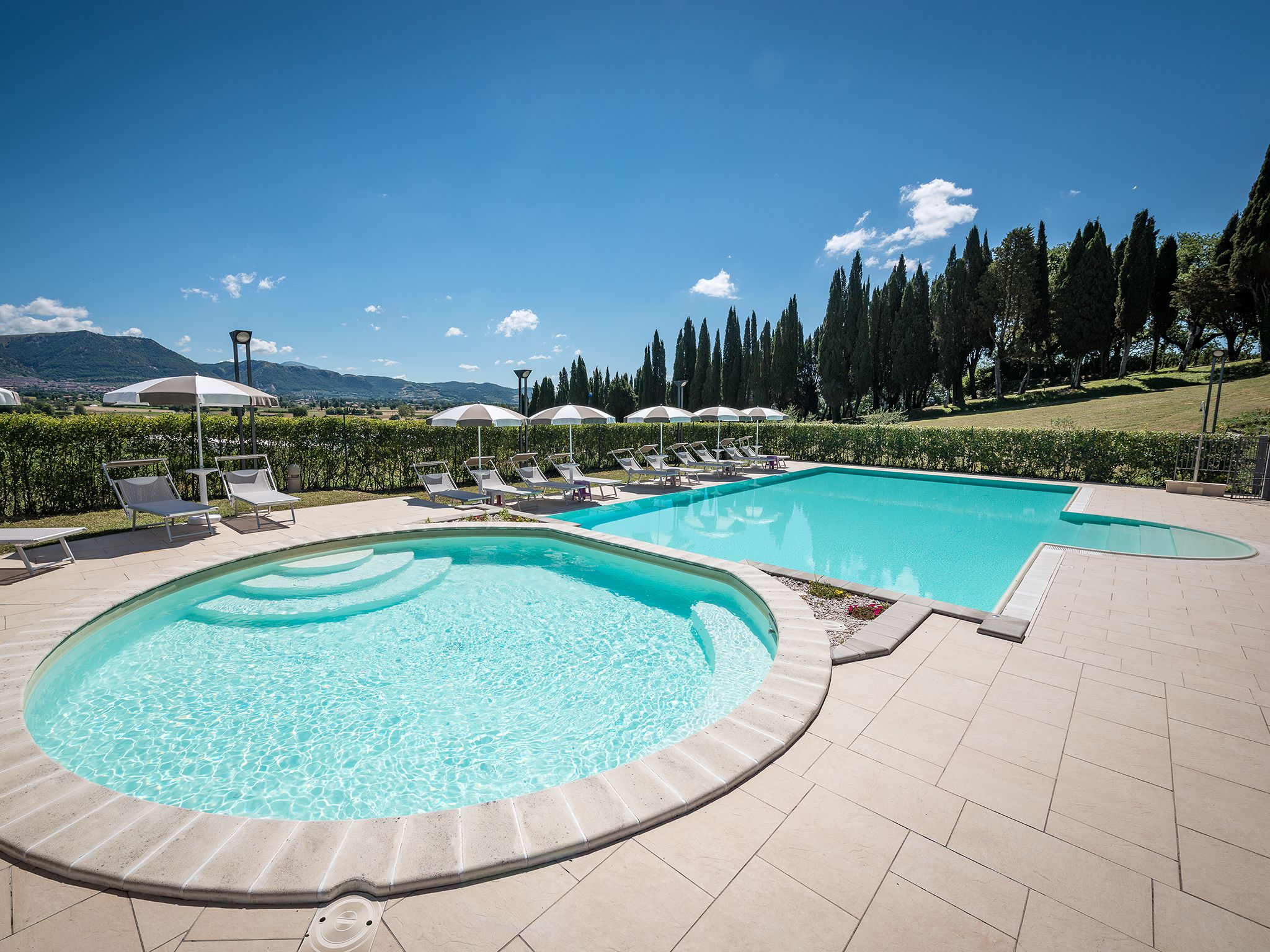 A swimming pool area with two pools, lounge chairs, and umbrellas.