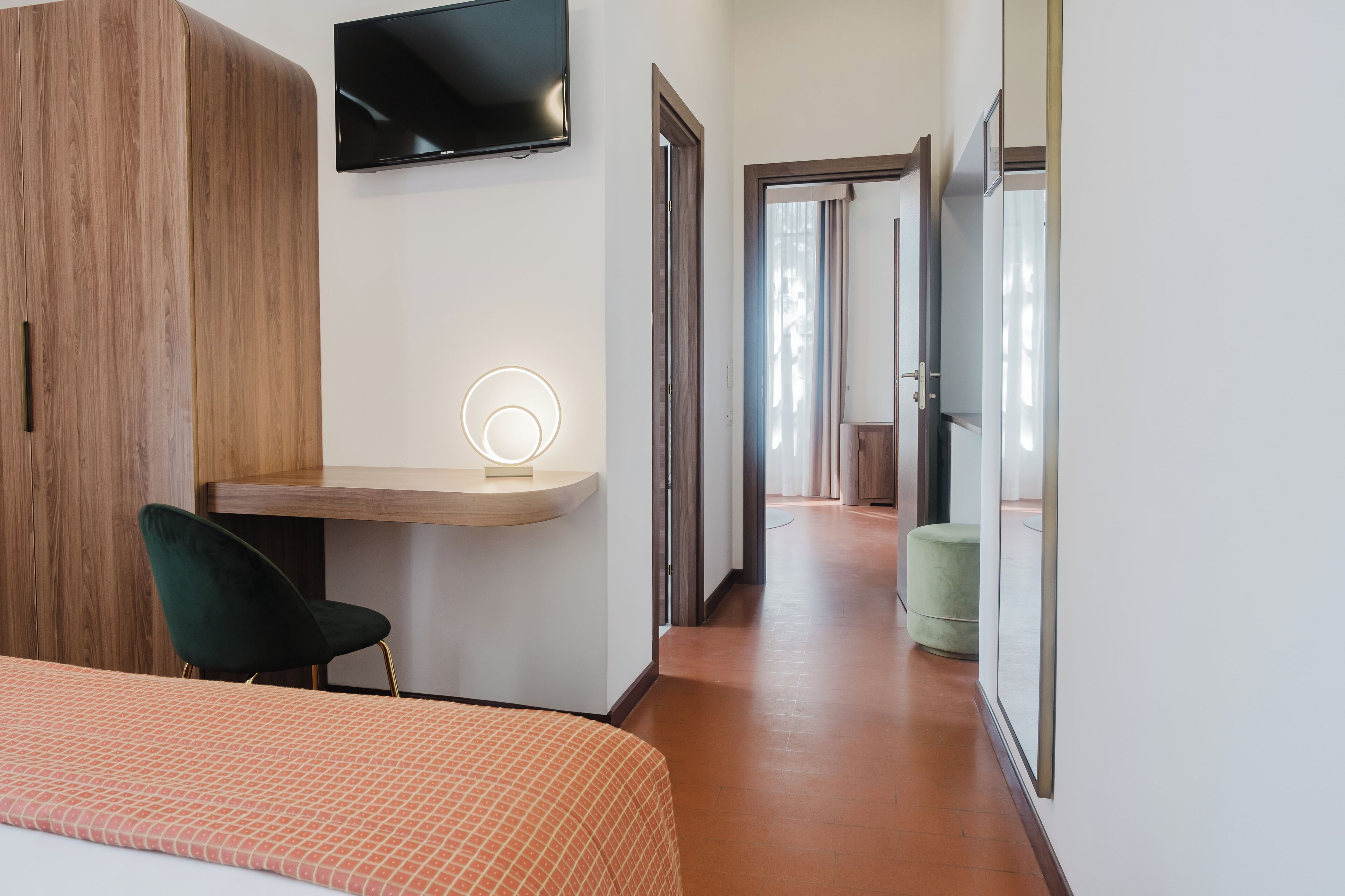 A hotel room with a bed, a desk with a chair, a TV mounted on the wall, and a hallway leading to another room.