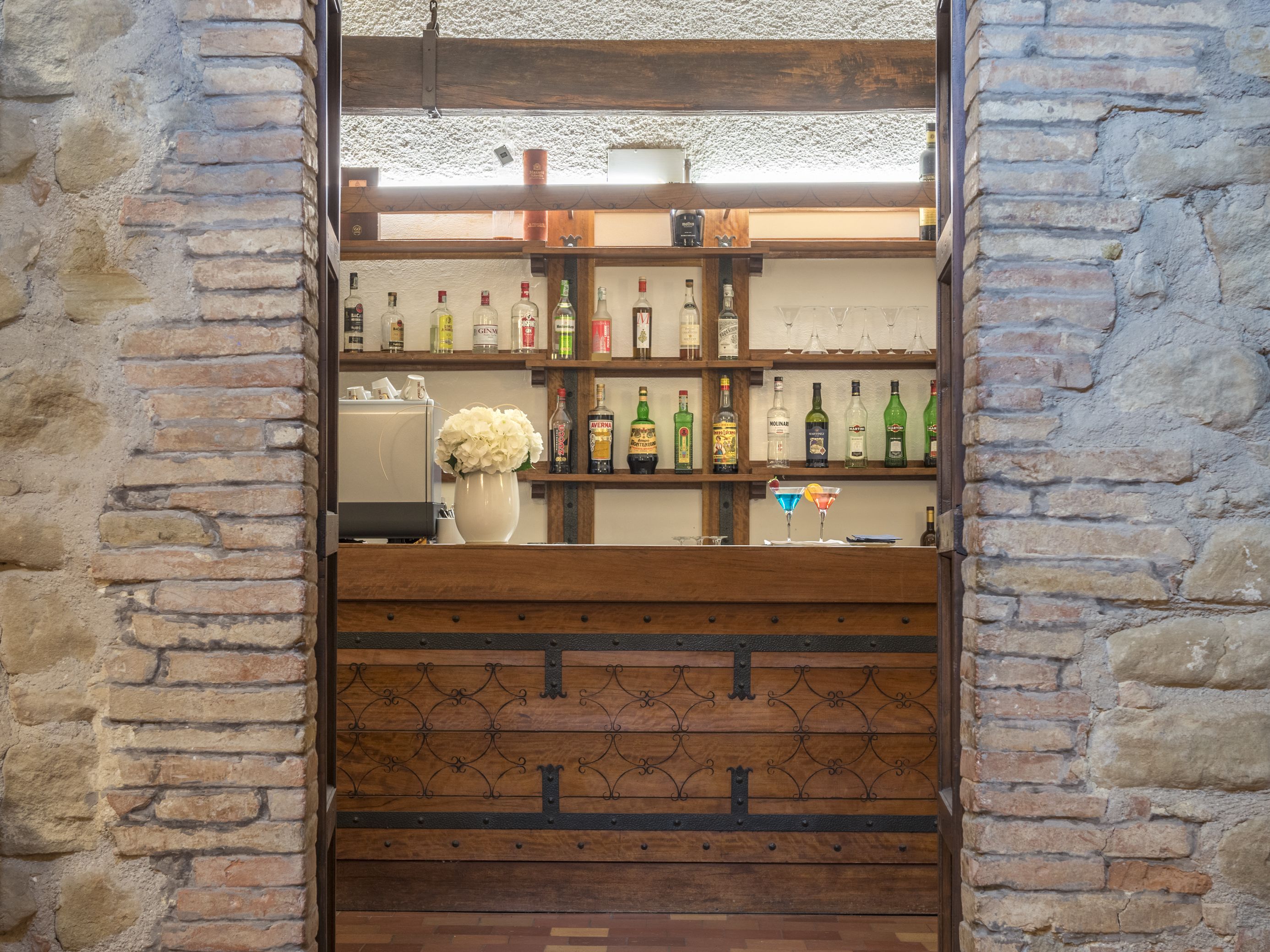 A bar area with shelves of liquor bottles, a white vase with flowers, and a wooden counter.