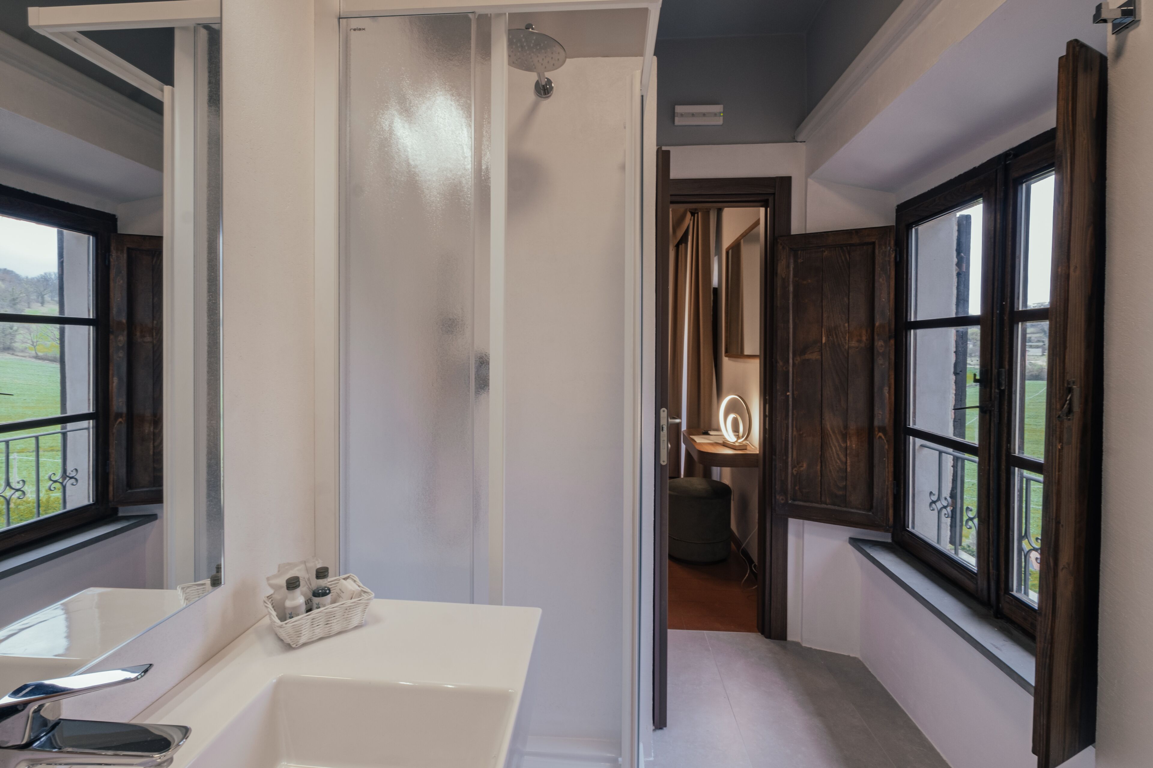 Un baño con bañera, ducha y tocador con espejo. Se ve un paisaje verde por la ventana.